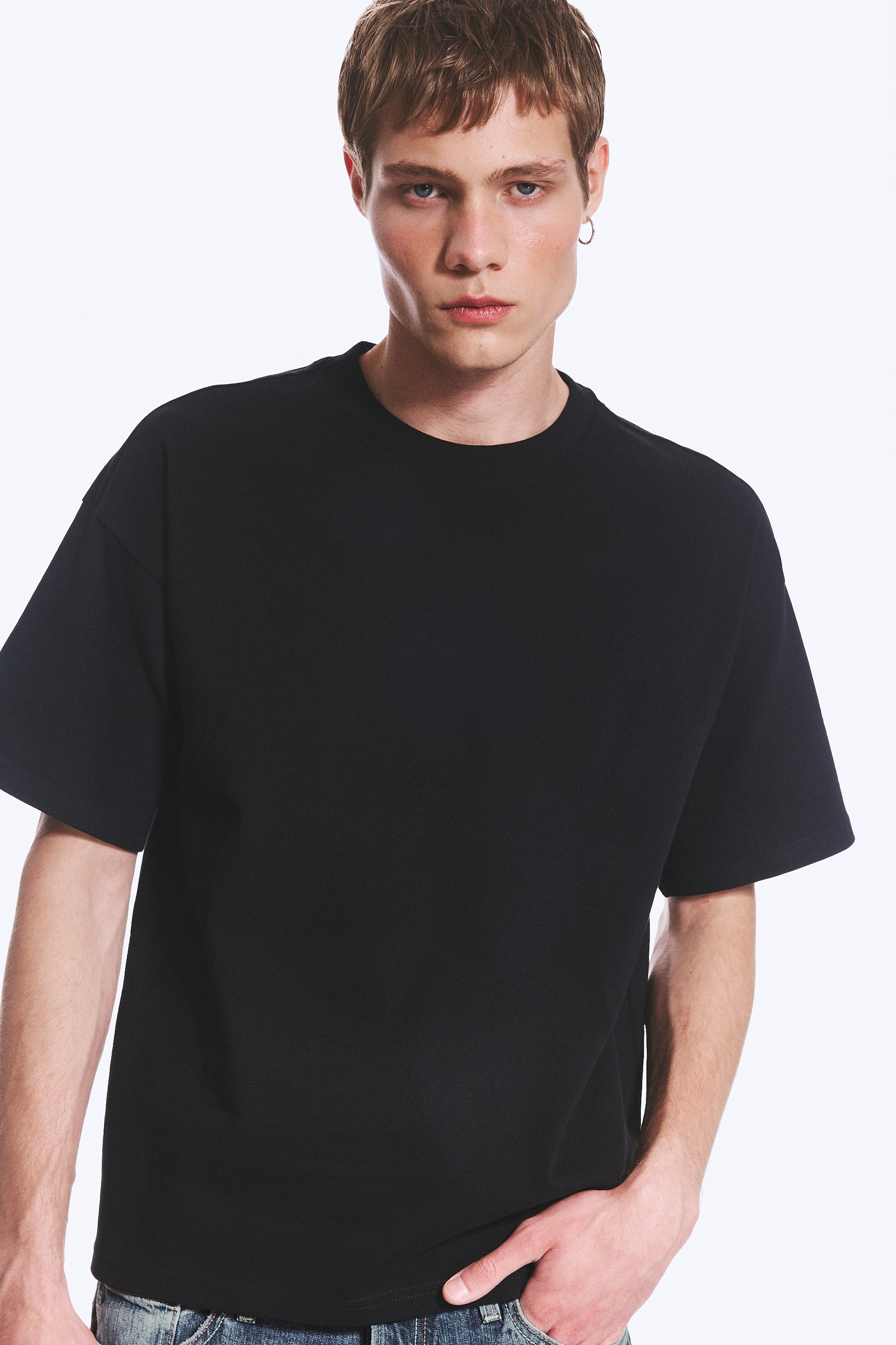 Ver imagem maior: Uma t-shirt preta oversized com gola redonda, ombros descaídos e mangas curtas largas está a ser usada pela modelo. A modelo está a colocar uma mão no bolso das calças de ganga, que são visíveis abaixo da t-shirt.