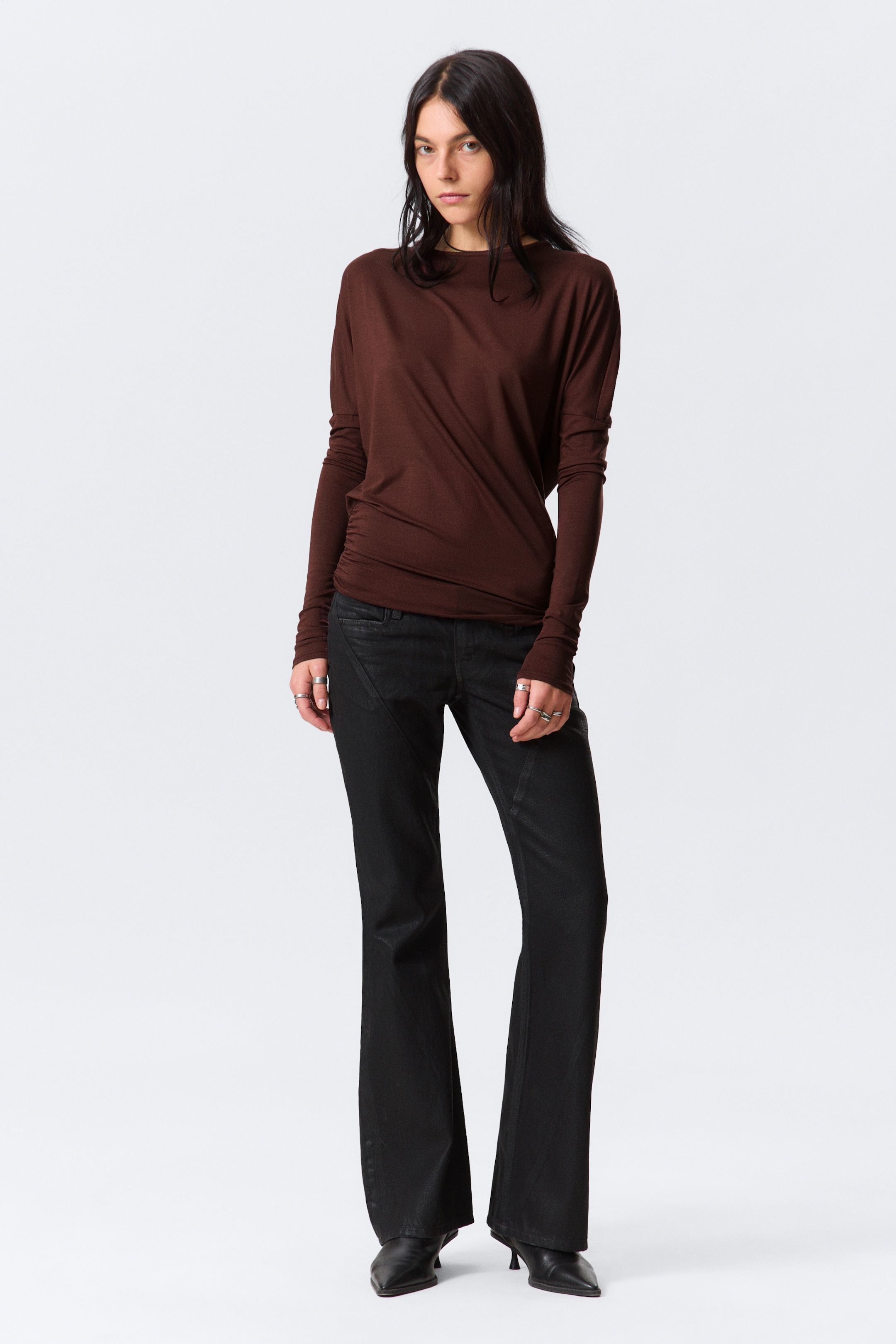 Agrandir l'image: Un haut à manches longues brun foncé, avec un large col rond, des poignets allongés et un design drapé et froncé sur le côté gauche du torse, est présenté. Un pantalon noir est porté en dessous, montrant une coupe ajustée en haut avec des détails de couture avant distincts et une légère évasée à l'ourlet.