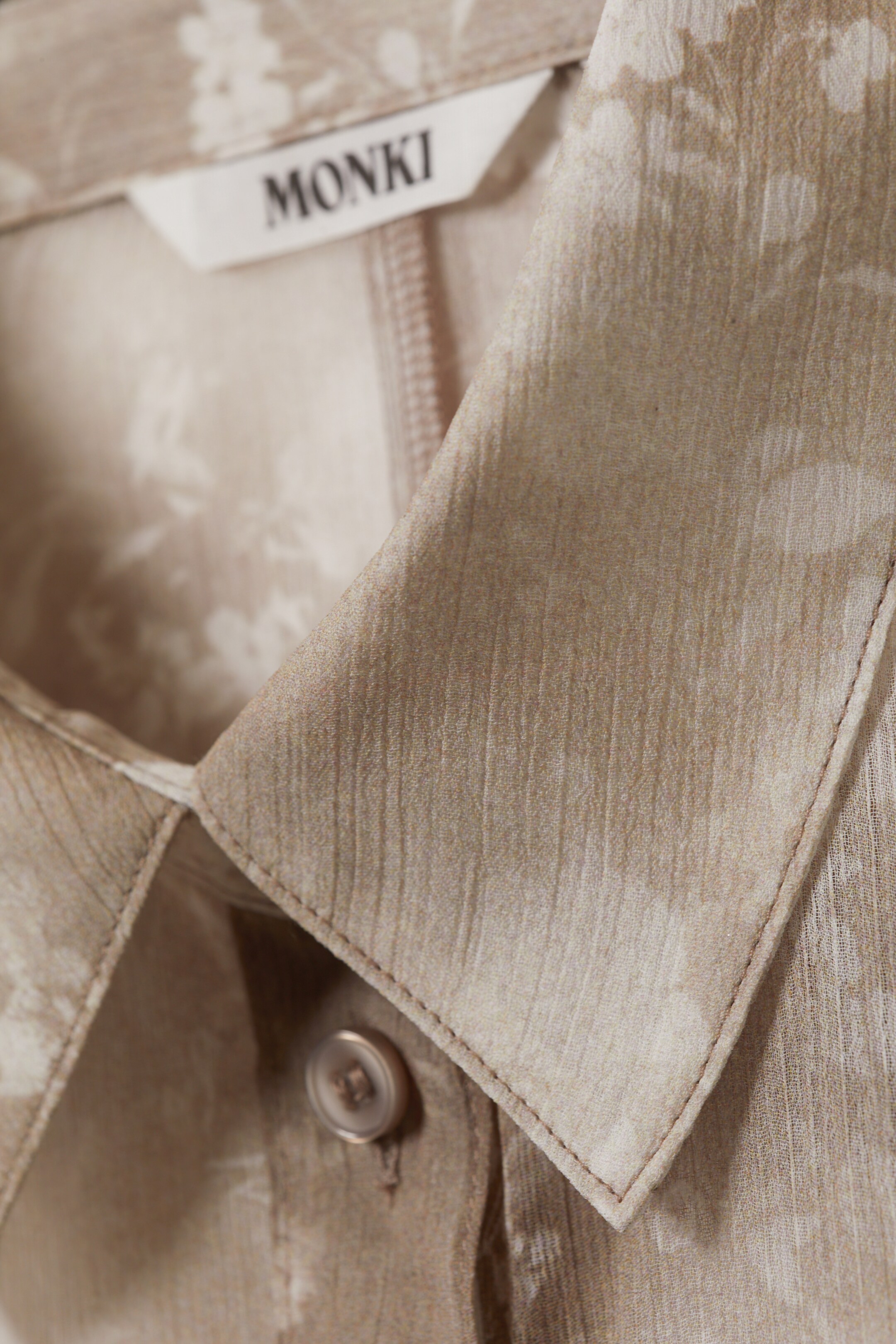 View larger image: A beige, crinkled shirt with a subtle floral pattern is displaying its classic pointed collar, neatly folded and showing visible stitching. A small, round button is fastened on the front placket, and a white "MONKI" brand tag is visible at the back of the neck, above a partially seen zipper.