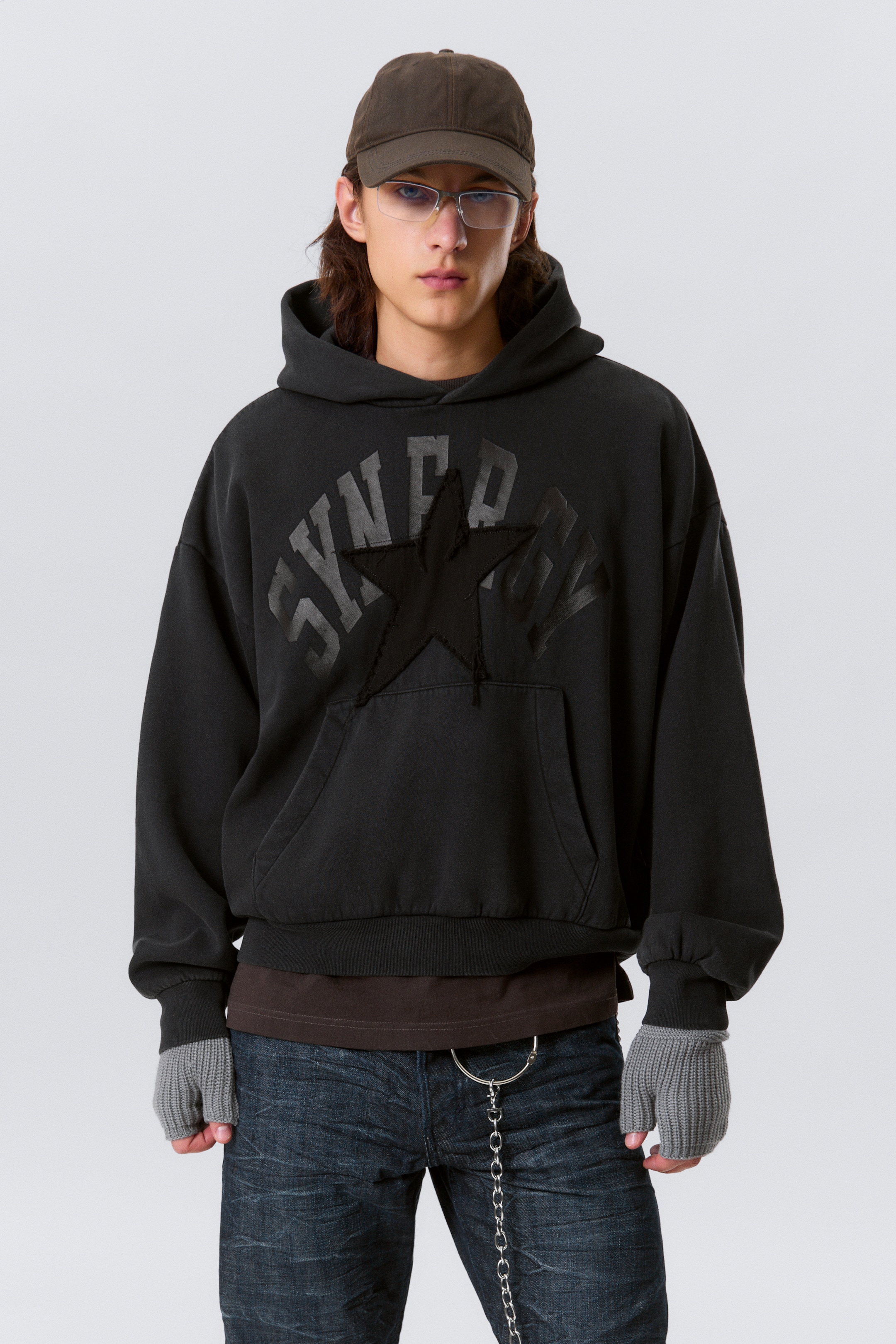 View larger image: A washed black oversized hoodie is being presented front-facing. It is featuring a large, distressed black star appliqué on the chest, which is centered above a spacious kangaroo pocket. Dark gray text is printed around the star, and the hoodie is designed with ribbed cuffs and hem, appearing slightly cropped over a brown t-shirt.
