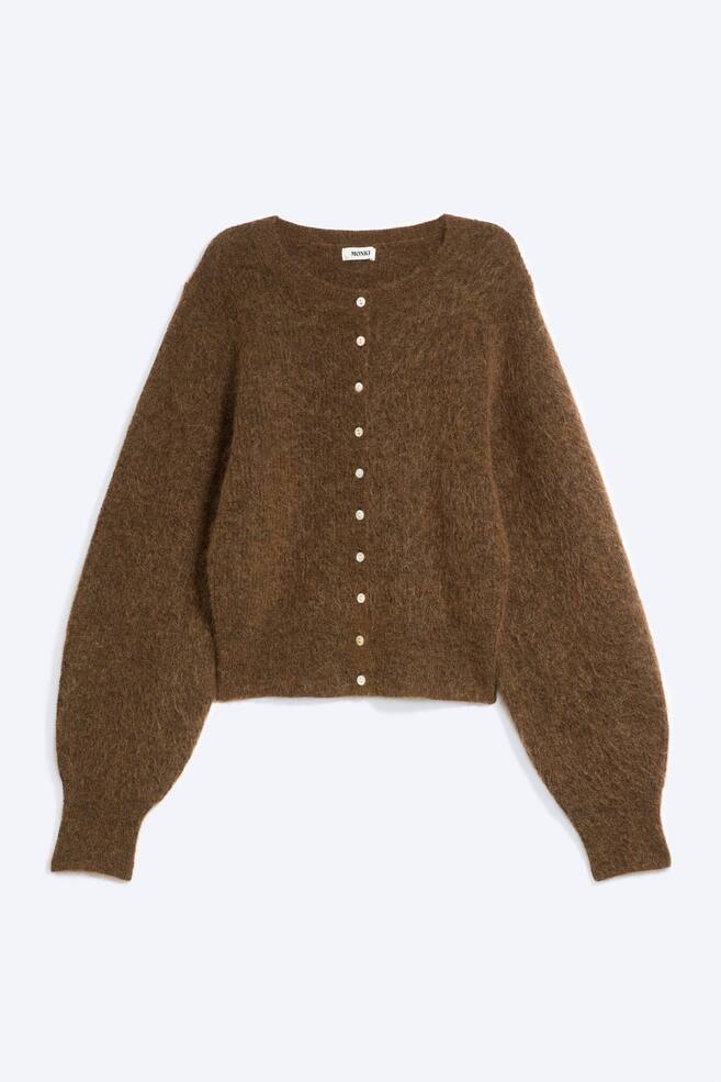 Een bruine cardigan in mohairmix wordt van voren getoond en heeft een zachte, geborstelde gebreide textuur, een ronde hals en een knoopsluiting aan de voorkant met kleine parelmoerachtige knopen. Het model heeft lange, volumineuze mouwen die samenkomen in brede, aansluitende ribboorden. - 1