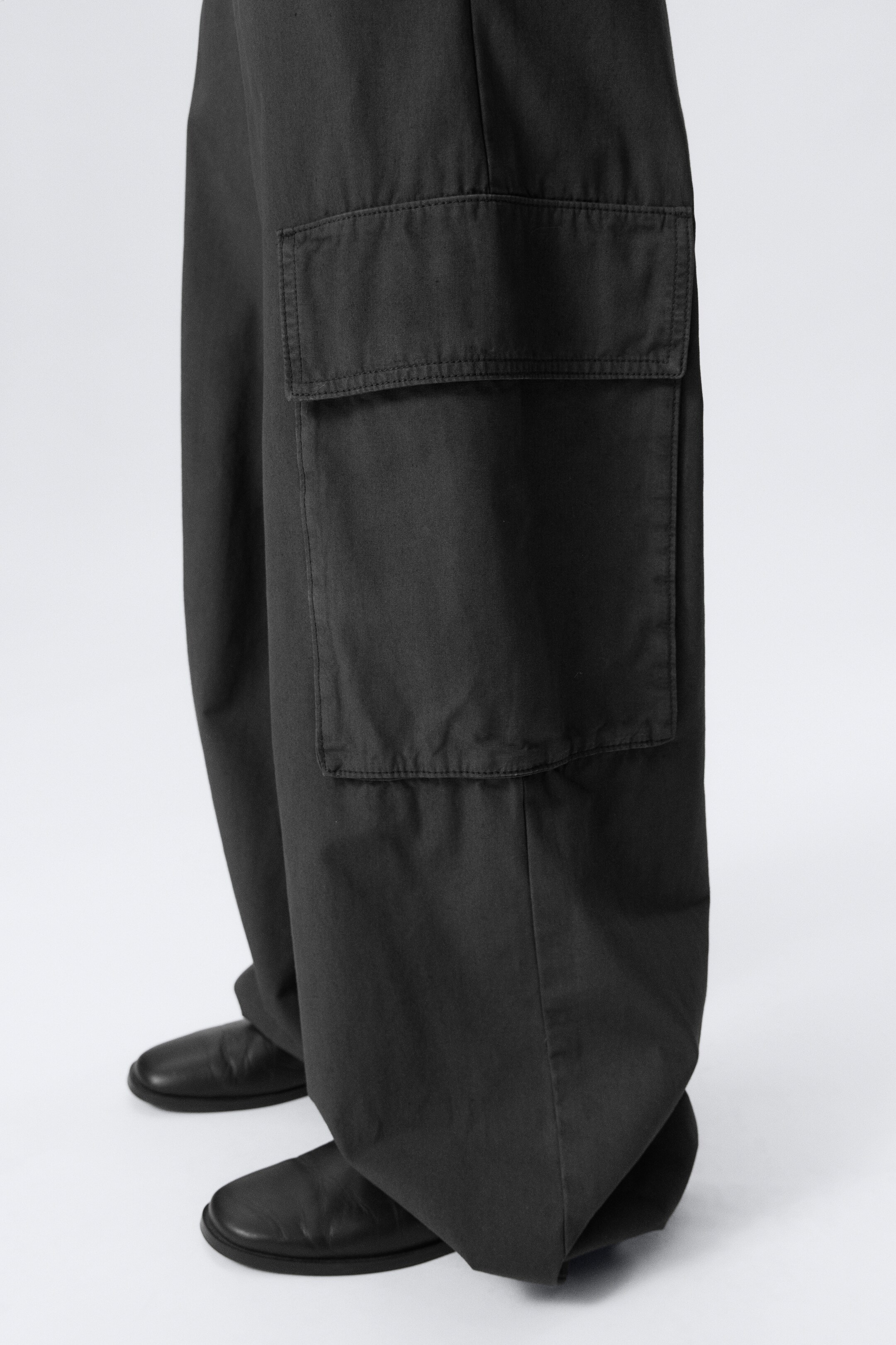 Vezi imaginea mai mare: Pantalonii cargo cu picior larg de culoare gri închis sunt purtați, având un buzunar mare cu clapetă pe partea laterală a piciorului. Țesătura se adună peste pantofii negri, subliniind croiala lejeră și relaxată.
