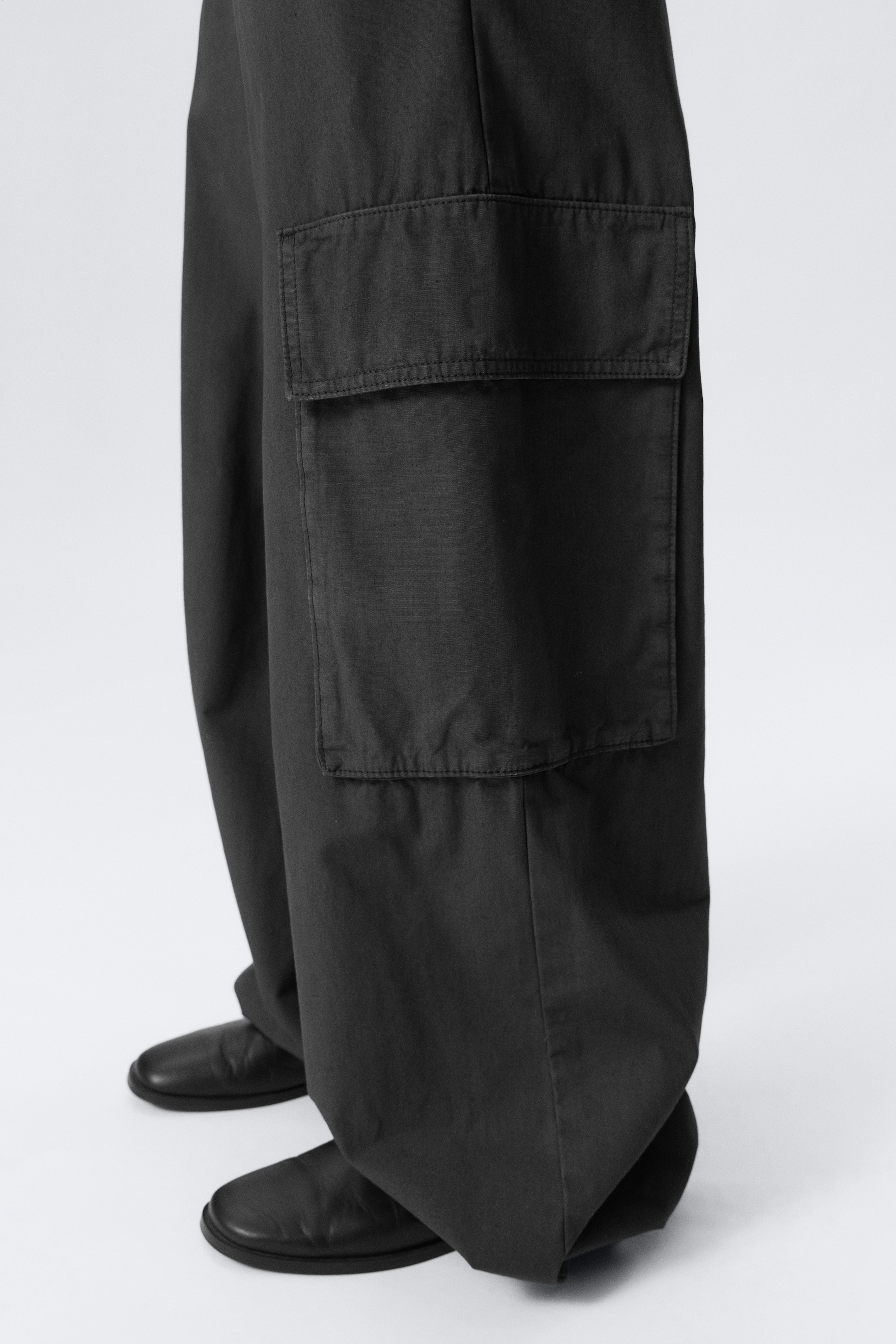 Größeres Bild ansehen: Dunkelgraue Cargo-Hosen mit weitem Bein werden getragen, mit einer großen Pattentasche an der Seite des Beins. Der Stoff sammelt sich über schwarzen Schuhen und betont den lockeren, entspannten Sitz.