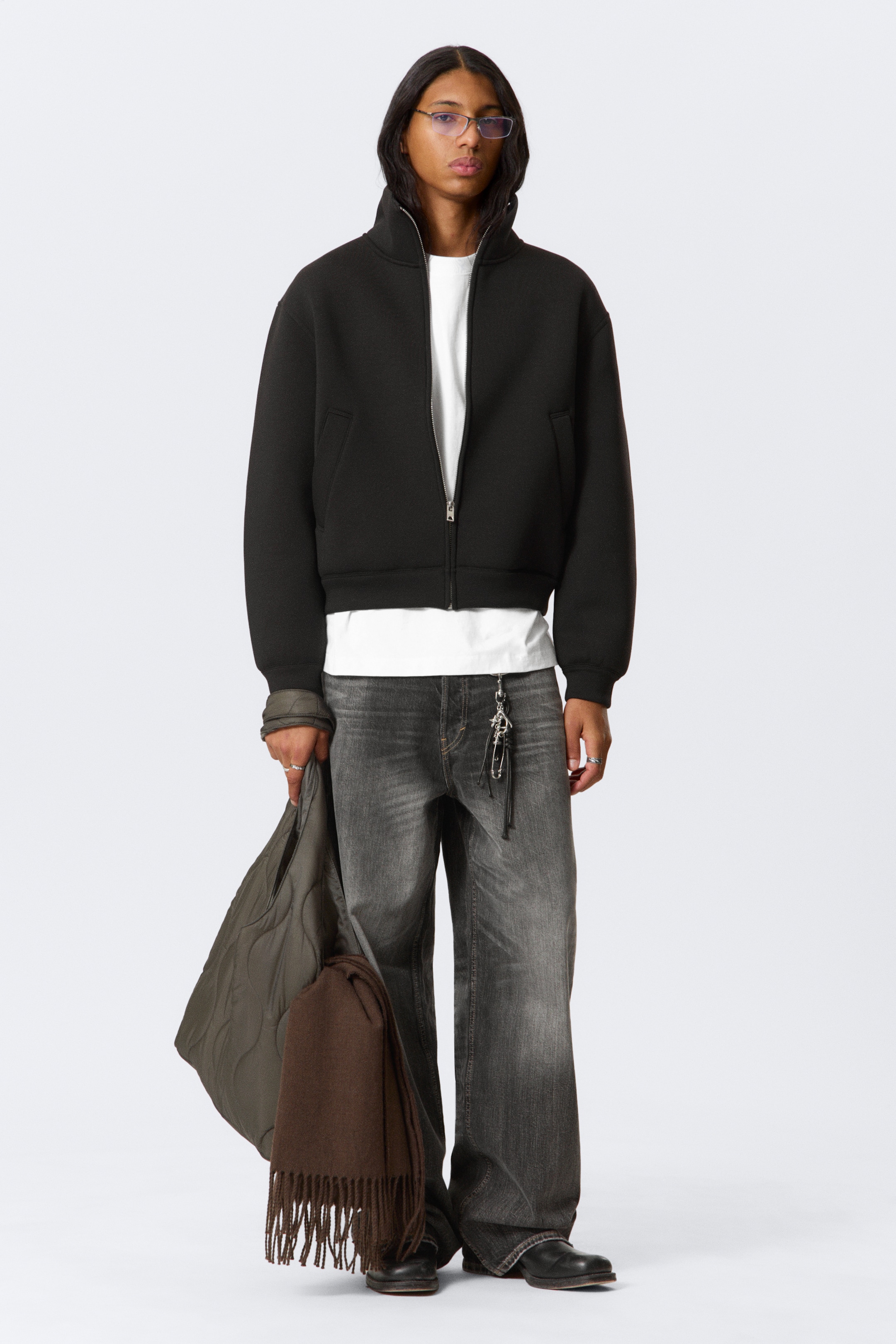 View larger image: A black bomber jacket is presenting a high stand collar and a full silver zip closure. It is appearing to be made of a thick, structured fabric, displaying horizontal welt pockets at the front, and is finished with ribbed cuffs and hem, creating a slightly cropped and relaxed silhouette.