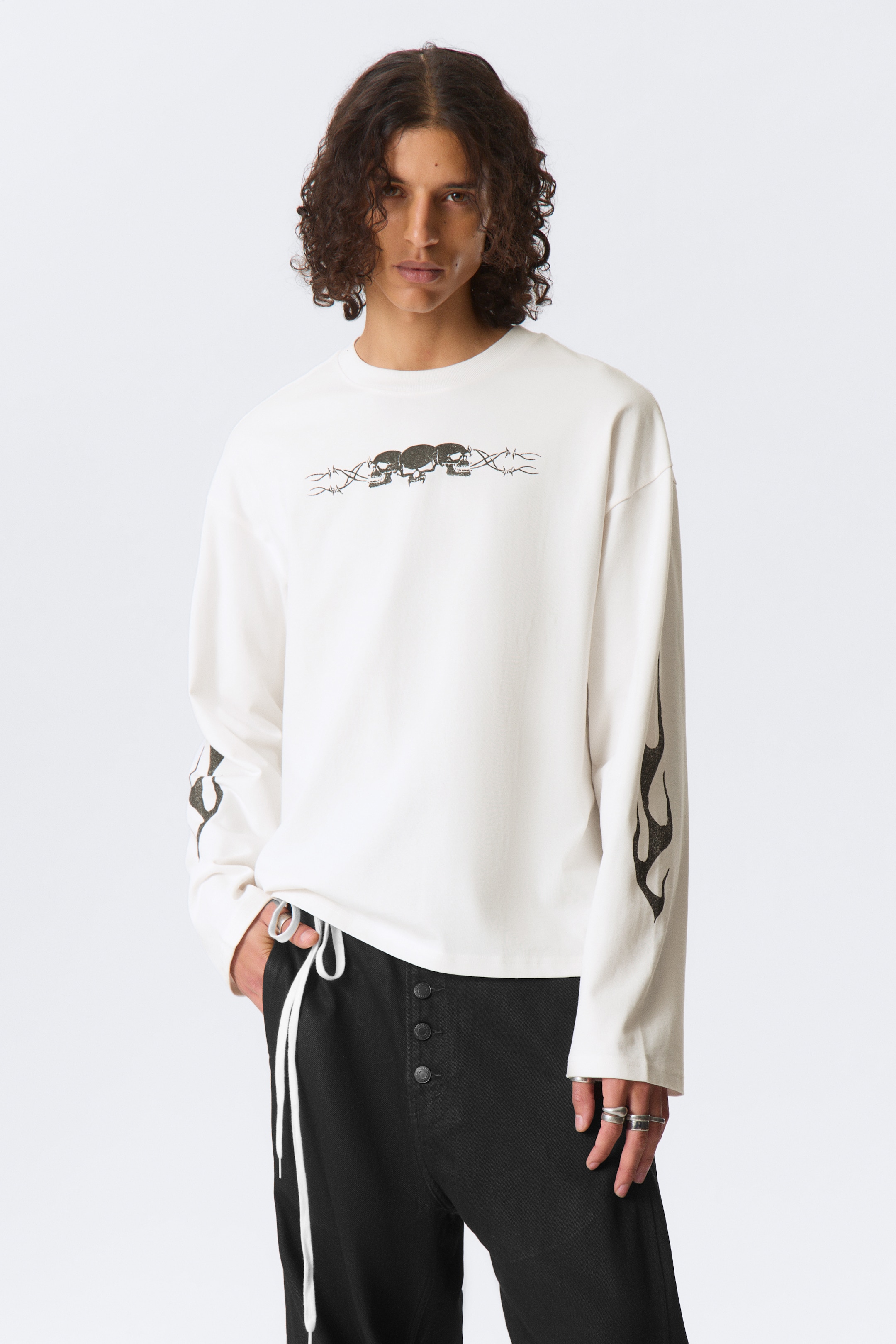 Vis større bilde: En off-white langermet skjorte har en sentral grafikk av tre mørke hodeskaller innrammet av piggtråd over brystet og mørke flammegrafikker på de ytre ermene. Skjorten har en avslappet passform med rund hals, vendt forover.