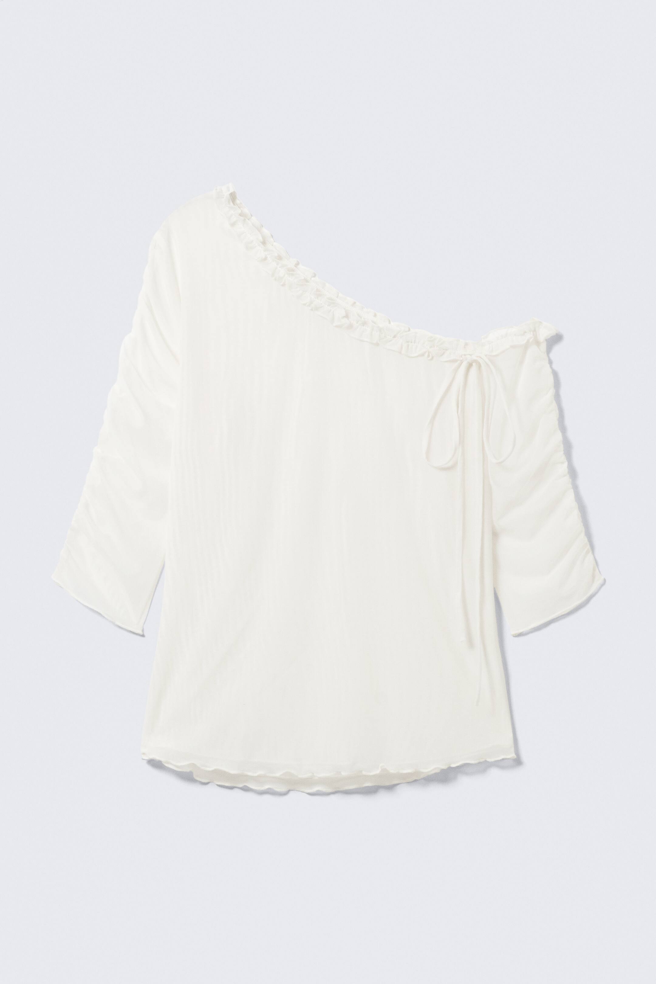 Näytä suurempi kuva: Valkoinen yksiolkaiminen toppi esittelee etupuoltaan, jossa on röyhelökoriste vasemmassa kaula-aukossa ja röyhelökoriste solmimisnauhoilla oikeassa olkaimessa. Molemmissa kolmivartisissa hihoissa on rypytysdetaljeja, ja helma on myös aaltoreunainen.