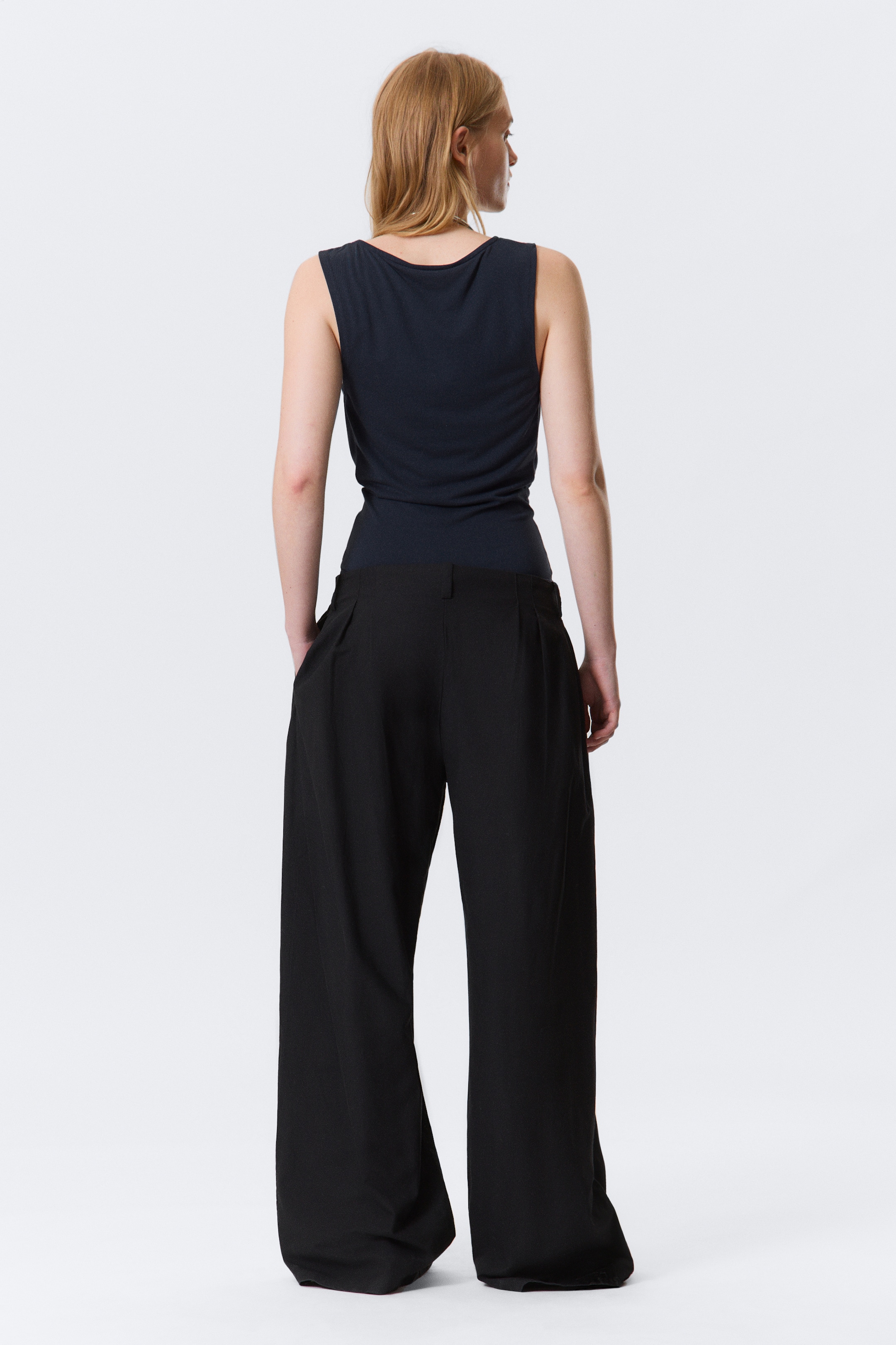 Ver imagen más grande: Una camiseta de tirantes oscura con escote redondo se lleva puesta, metida dentro de unos pantalones negros de pierna ancha con pliegues traseros visibles, mientras la persona está de espaldas.