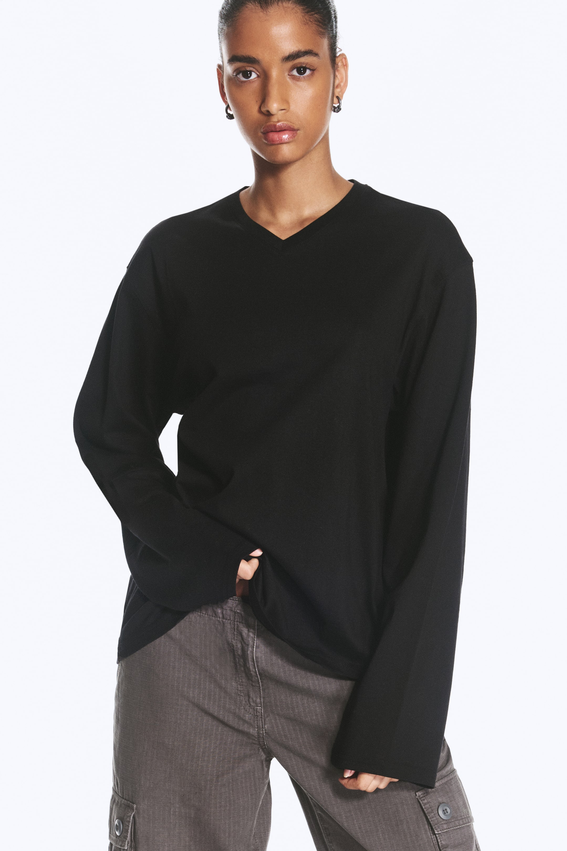 Grotere afbeelding bekijken: Het model draagt een zwart, boxy T-shirt met V-hals en lange mouwen, met een ontspannen pasvorm en wijde mouwen. Het T-shirt is gestyled met grijze cargobroeken, waarbij het model de zoom iets bij de taille vasthoudt.