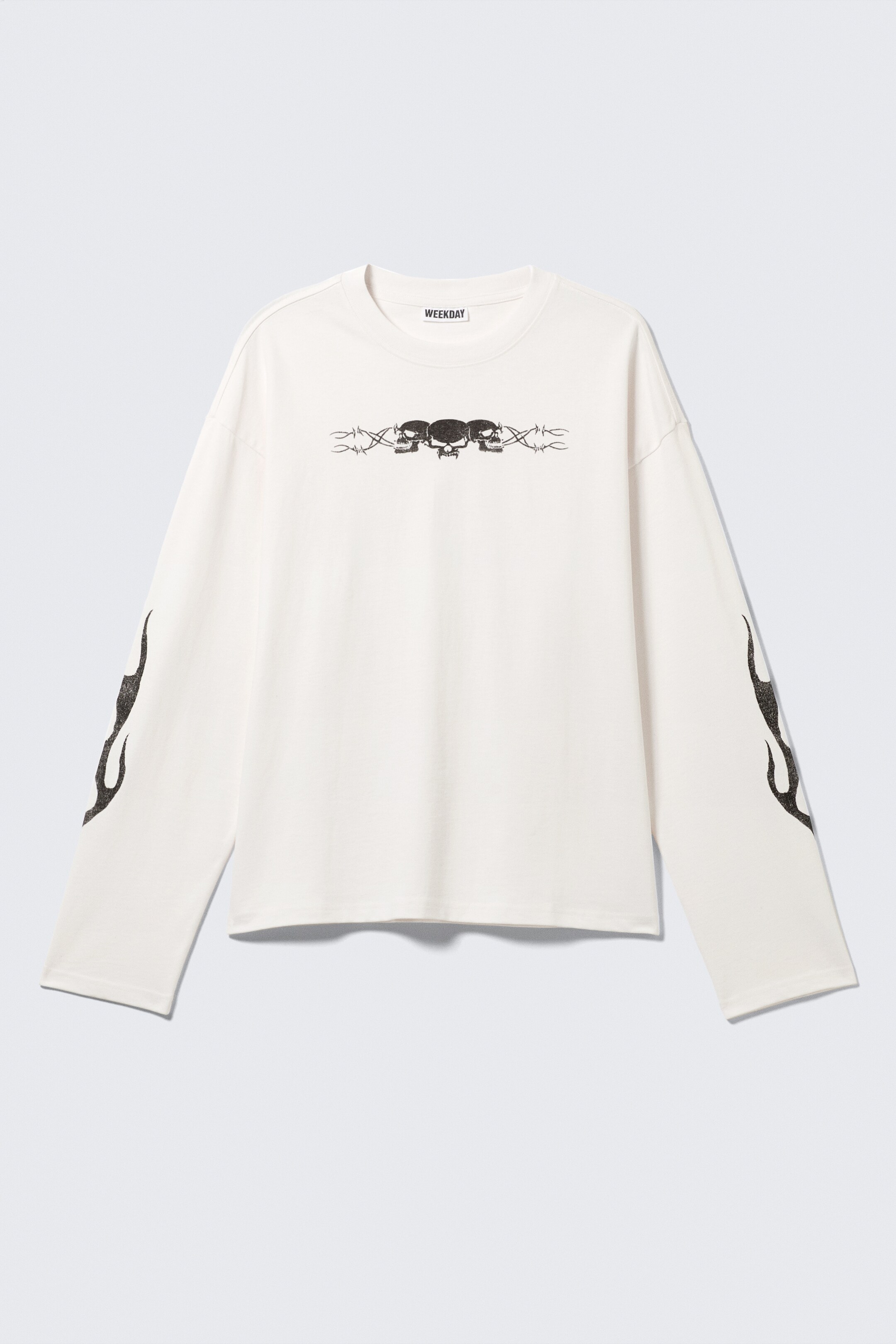 Ingrandisci l'immagine: Una T-shirt a maniche lunghe, di colore bianco sporco e dalla linea squadrata, è mostrata davanti, caratterizzata da uno scollo rotondo a coste, orli a doppia cucitura e un'etichetta del marchio 'WEEKDAY' sul retro del collo. Un grafico di tre teschi con filo spinato è centrato sul petto, e delle grafiche di fiamme compaiono sui lati esterni di entrambe le maniche.