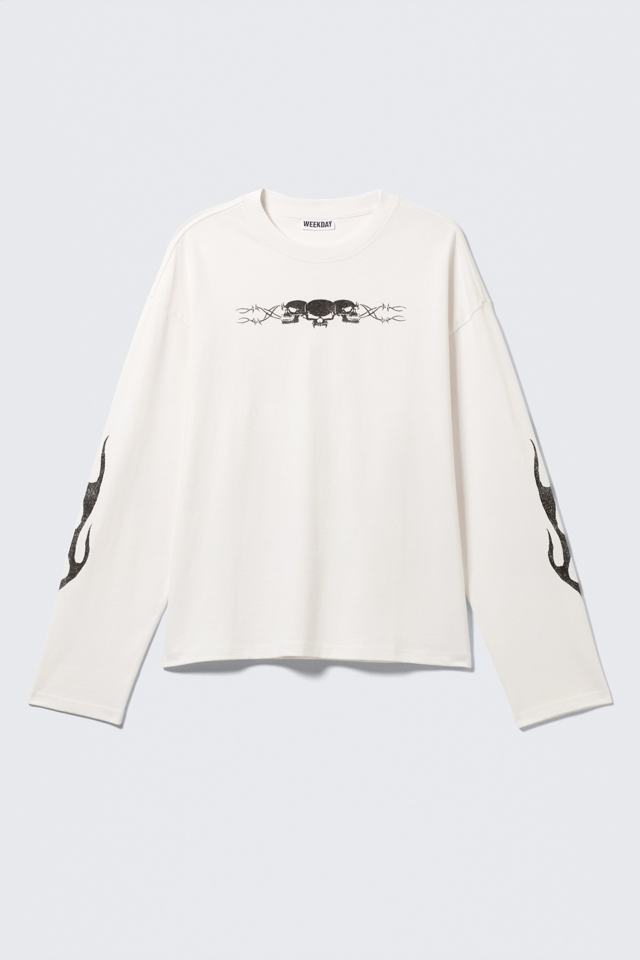 Visualizza immagine più grande: Una T-shirt color avorio, squadrata, a maniche lunghe è mostrata dalla parte anteriore, con un collo rotondo a coste, orli a doppia cucitura e un'etichetta del marchio 'WEEKDAY' sul retro del collo. Un'immagine grafica di tre teschi con filo spinato è centrata sul petto e grafiche di fiamme appaiono sui lati esterni di entrambe le maniche.