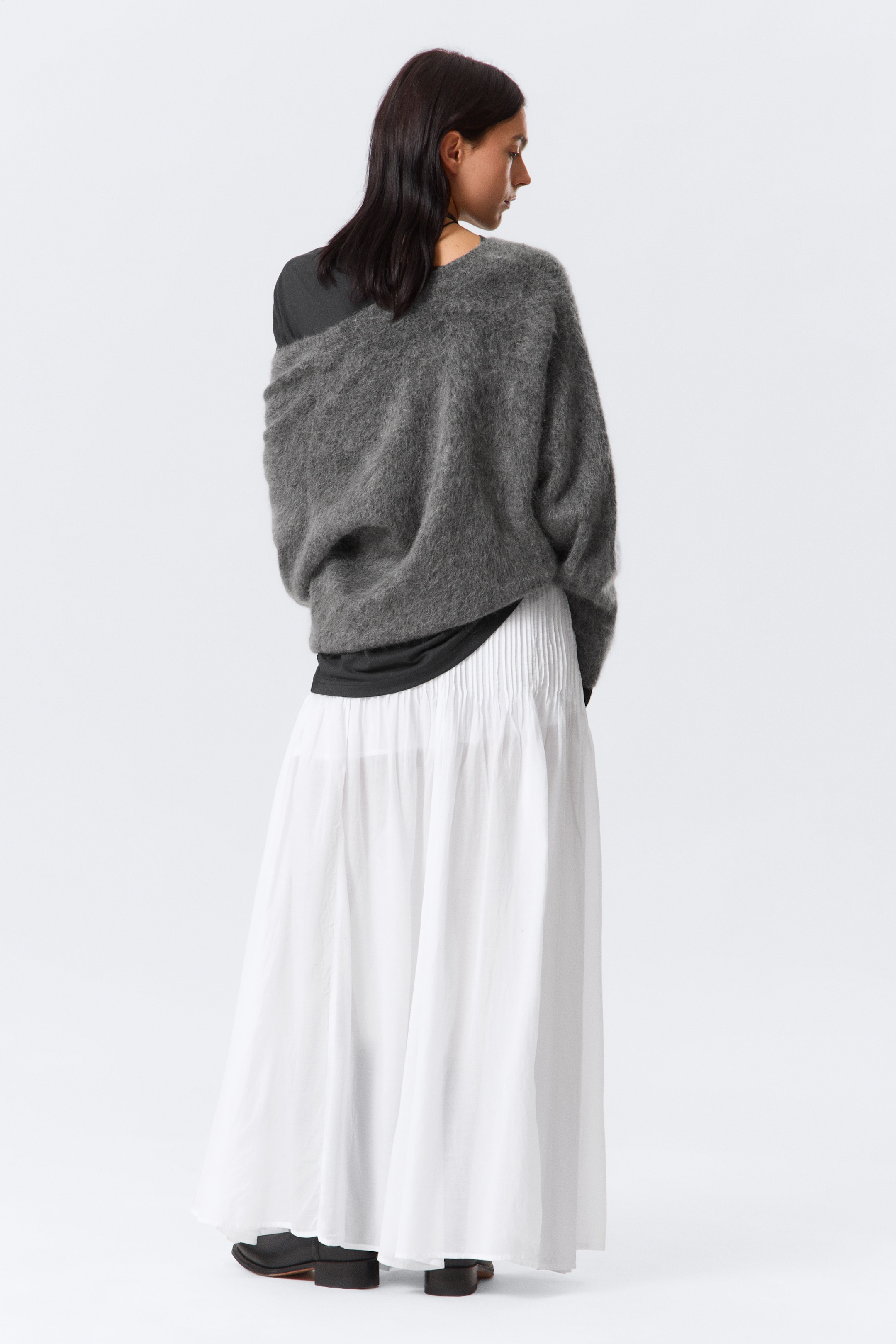 Agrandir l'image: Un pull gris, duveteux et tricoté, de grande taille est porté, tombant d'une épaule et superposé à un haut sombre, vu de dos. Il est associé à une jupe blanche, longue, avec des plis fins ou des fronces à la taille.