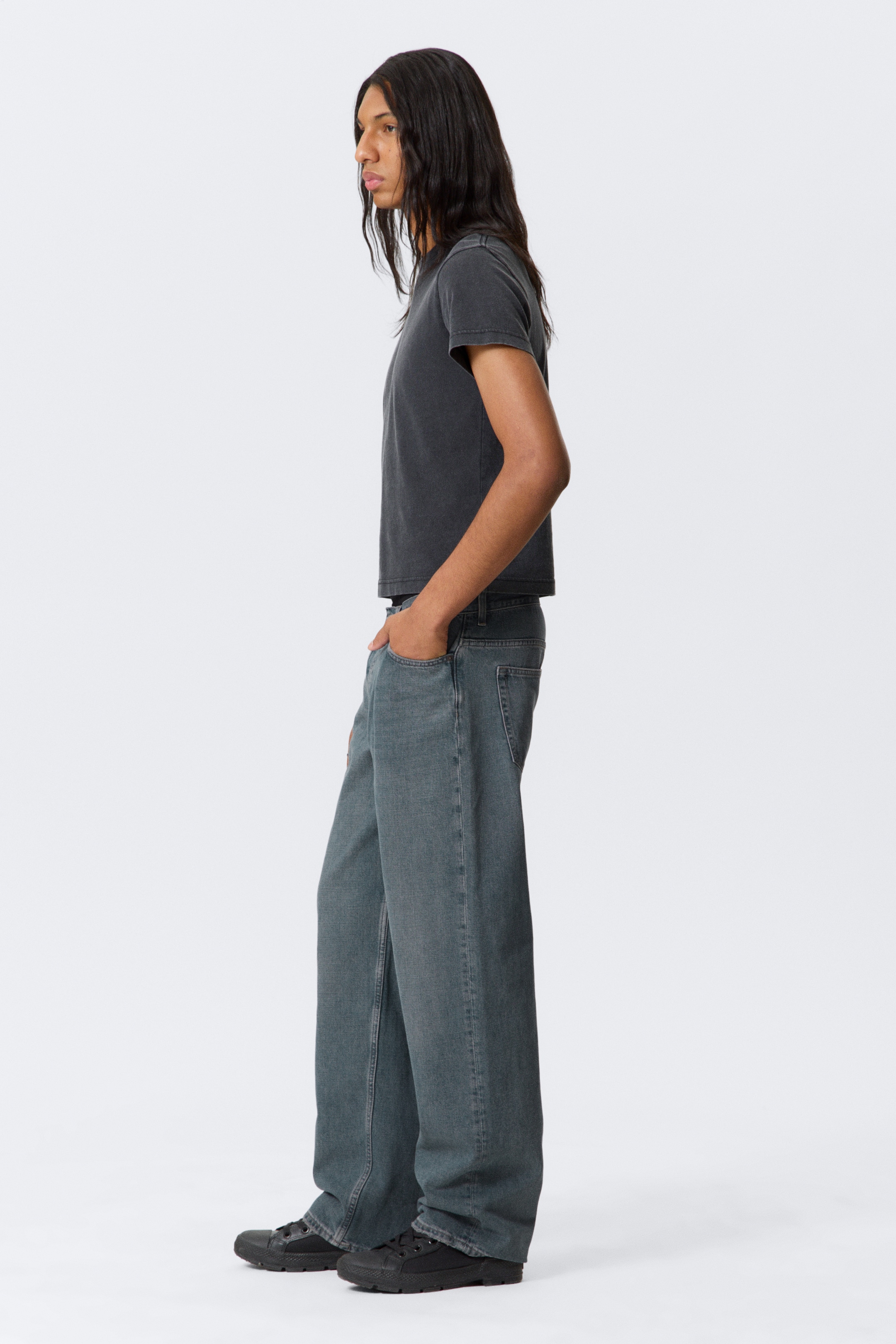 Ingrandisci l'immagine: Una maglietta a maniche corte di colore grigio scuro leggermente sbiadito, con scollo a girocollo, è indossata insieme a jeans a gamba larga, blu-grigio sbiaditi e a vestibilità rilassata. I jeans coprono visibilmente delle sneakers nere a collo basso, con suole spesse, il tutto mostrato da un profilo laterale sinistro.