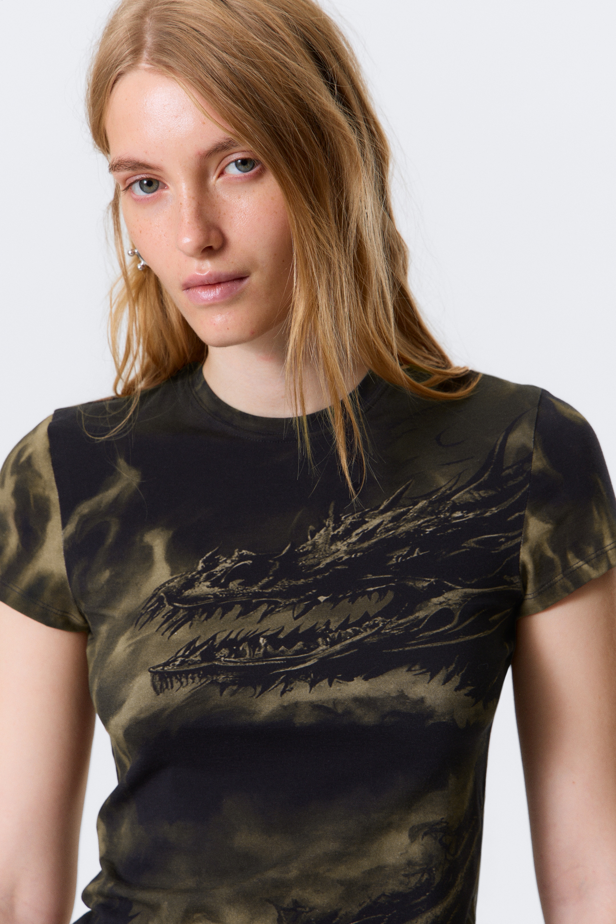 Größeres Bild anzeigen: Ein dunkles, eng anliegendes T-Shirt mit Rundhalsausschnitt und kurzen Ärmeln wird aus einer frontalen Perspektive präsentiert. Es zeigt ein großes Grafikdesign von zwei olivfarbenen Drachenköpfen auf der Vorderseite, die vor einem schwarz-olivgrünen Batikmuster gesetzt sind.