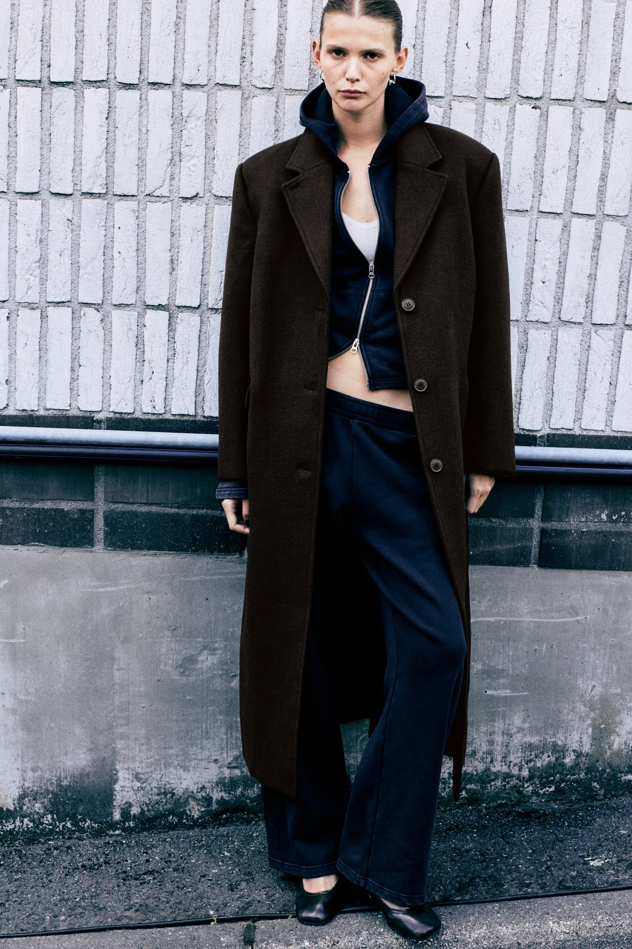 Se større billede: En oversized, lang mørkebrun car coat bæres åbent af modellen og har markerede skuldre, revers med hak og knapdetaljer foran. Frakken er stylet over en mørkeblå hoodie med lynlås og matchende sweatpants.