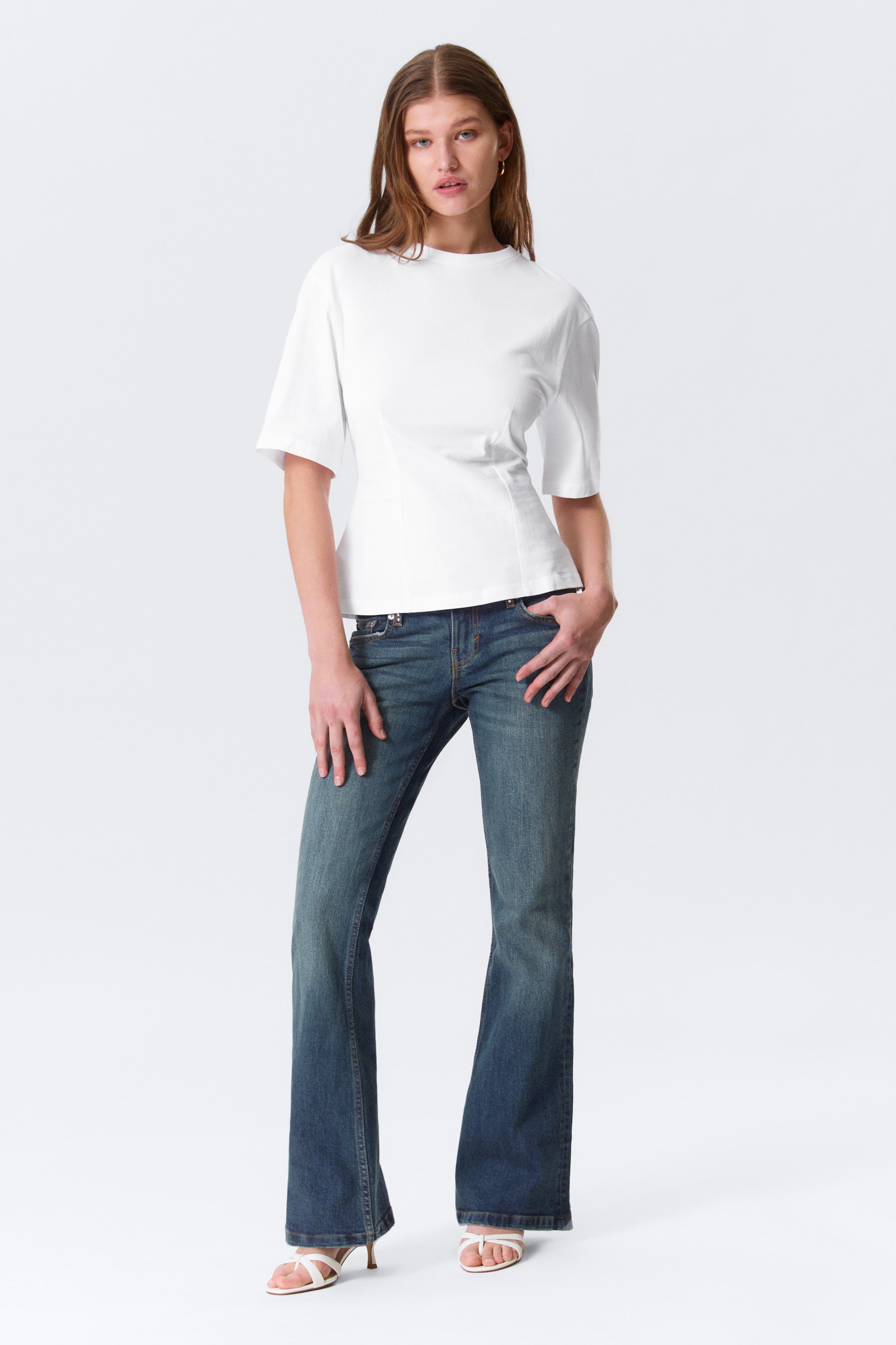 Grotere afbeelding bekijken: Een witte aansluitende top met een ronde hals, wijde ellebooglange mouwen en verticale naden die de taille definiëren, gecombineerd met medium-wash, laagzittende bootcut jeans, alles gepresenteerd in een vooraanzicht.