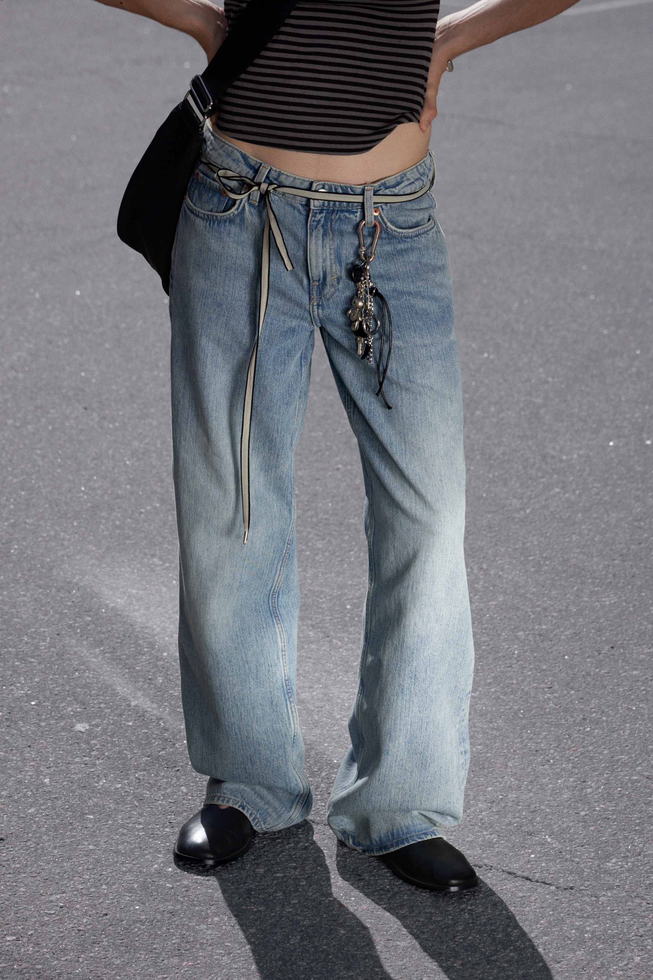 Größeres Bild anzeigen: Ein Paar hell gewaschene Jeans wird gezeigt, mit einem lockeren, weit geschnittenen Bein, das zu den unteren Beinen hin verblasst. Ein dünner, gestreifter Bandgürtel wird vorne in der Taille zu einer Schleife gebunden, wobei ein Ende lang herunterhängt, und ein Bündel verschiedener Schlüsselanhänger hängt von einer vorderen Gürtelschlaufe, während die Jeans über schwarzen Schuhen zusammenfällt.