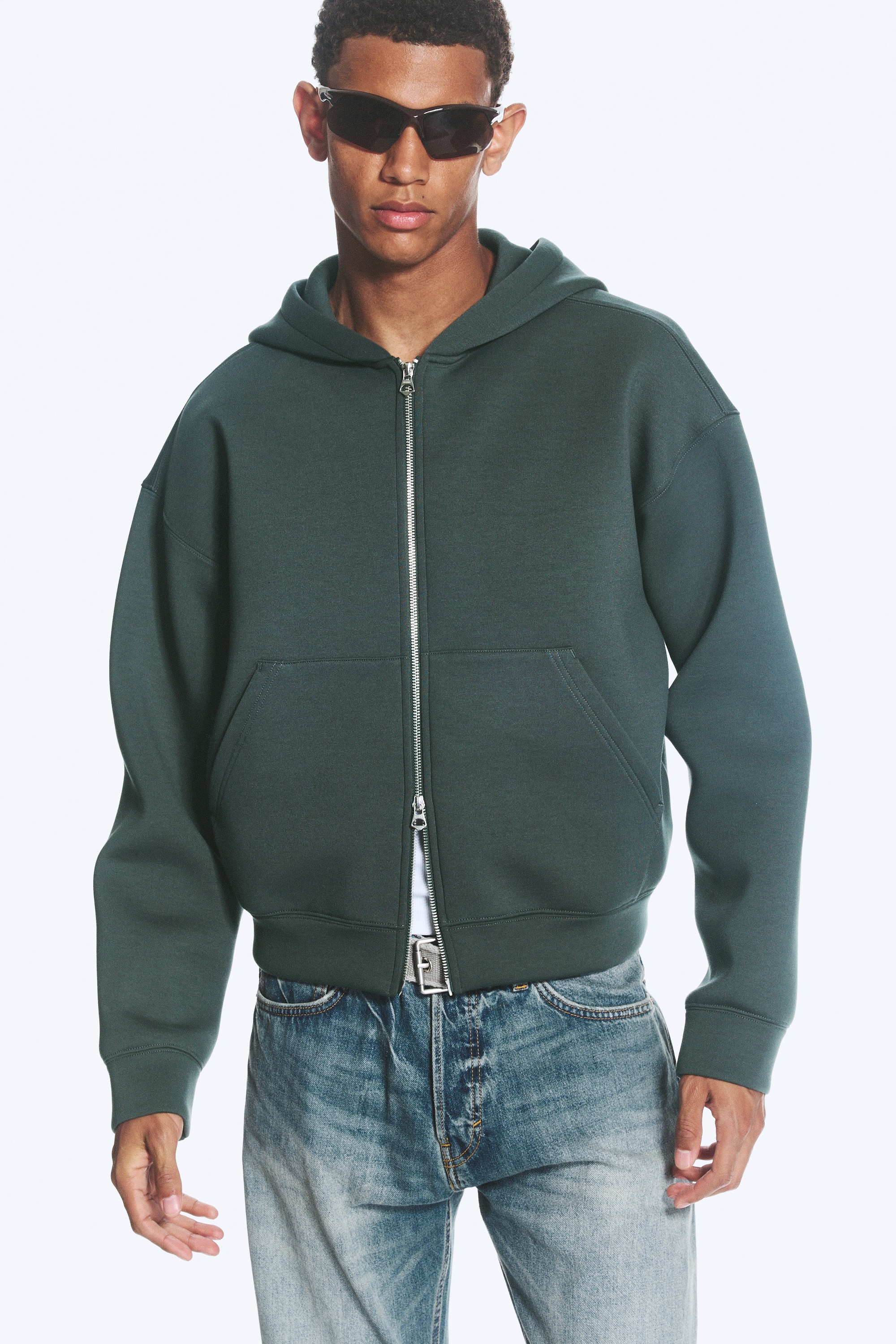 Um casaco com capuz verde escuro com fecho de correr está exibido numa vista frontal, deitado plano. Apresenta um fecho de correr prateado de comprimento total, dois bolsos frontais espaçosos e um capuz acoplado com uma etiqueta de marca "WEEKDAY" visível.