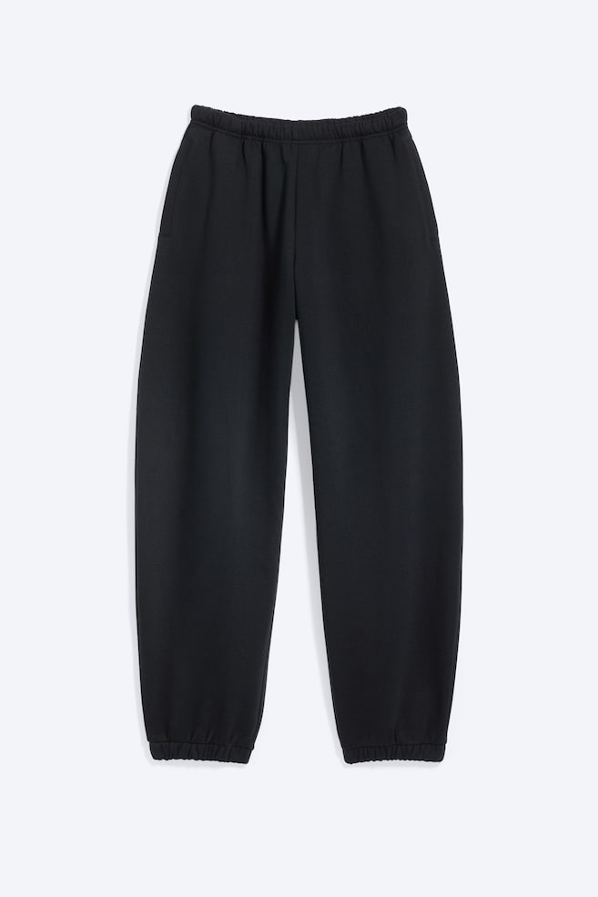 Los pantalones de chándal negros se muestran en vista frontal, con una cintura elástica, bolsillos laterales de ojal y piernas holgadas que se estrechan hacia los puños elásticos. - 1