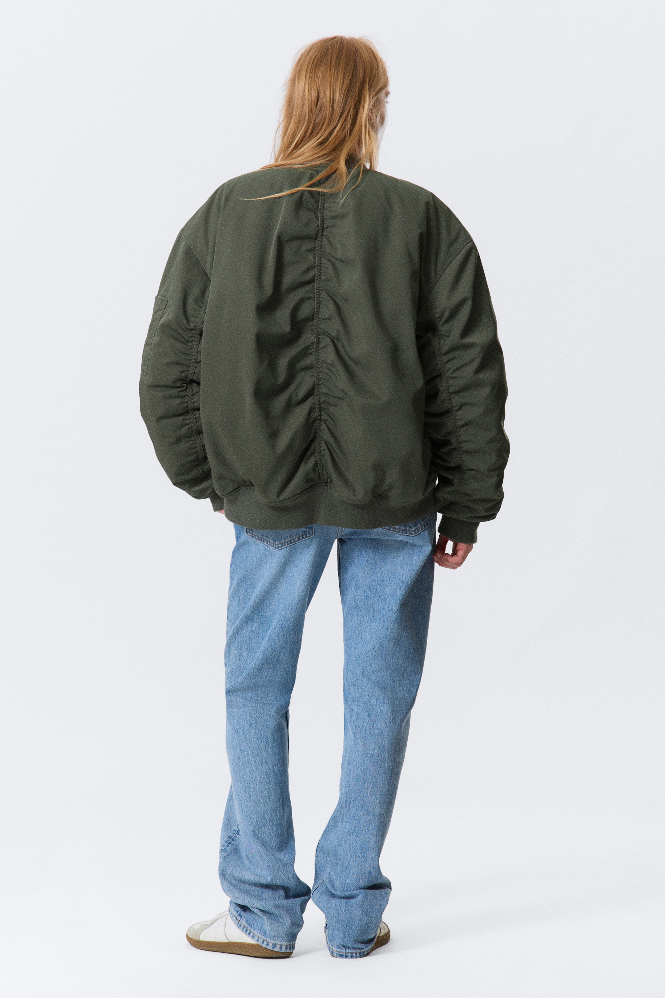 Visualizza immagine più grande: Un bomber verde oliva viene visto da dietro, mettendo in mostra la sua vestibilità oversize. Presenta dettagli raccolti verticalmente lungo il centro della schiena e arricciature orizzontali sulle maniche lunghe, che sono rifinite con polsini a coste e un orlo a coste.
