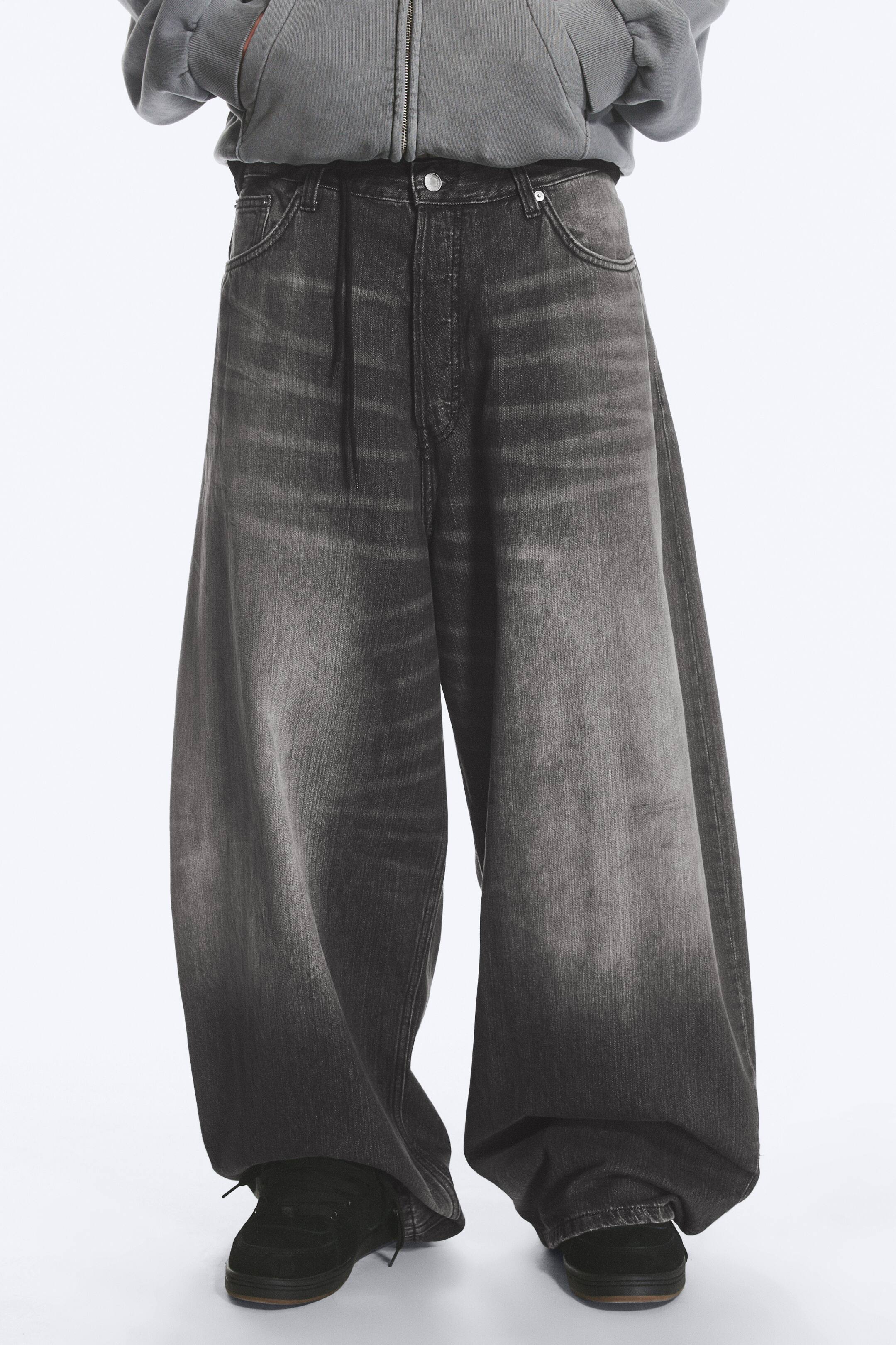 Grösseres Bild ansehen: Dunkelgraue Jeans mit weitem Bein, die mit einer ausgewaschenen Optik, einem Knopfverschluss, einem sichtbaren Kordelzug am vorderen Bund und Vordertaschen ausgestattet sind. Die Jeans präsentieren einen extrem weiten Schnitt, der sich über dunklen Schuhen an den Knöcheln sammelt.