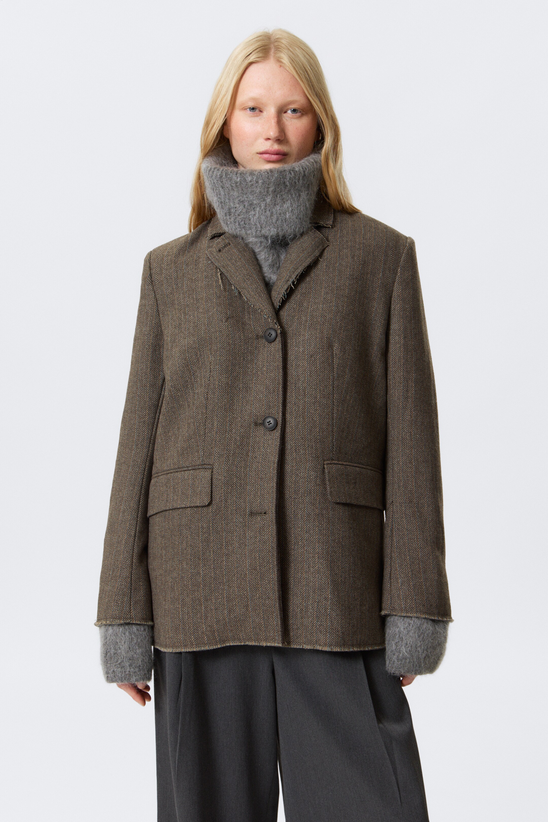 Agrandir l'image: Un blazer marron à chevrons à simple boutonnage, avec des revers crantés, des poignets et un ourlet avec des détails de bord brut, ainsi que des poches à rabat à l'avant, est porté par le mannequin. Il est superposé à un col roulé en grosse maille grise et un pantalon gris foncé.