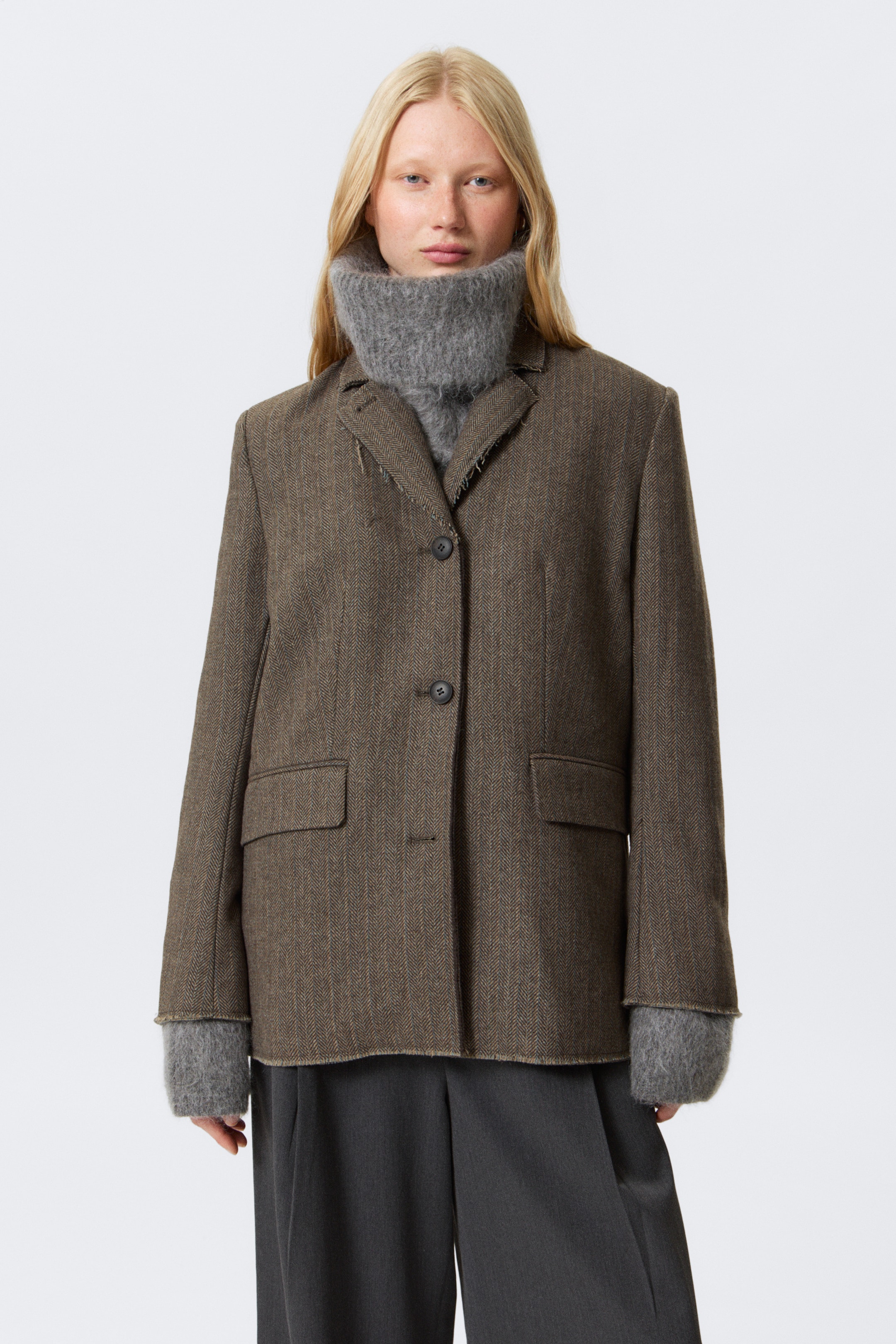 Vis større bilde: En brun fiskebensmønstret enkelspent blazer, med hakkede slag, mansjetter og en kant med rå kantdetaljer, sammen med frontlommer med klaff, bæres av modellen. Den er lagdelt over en grå chunky strikket høyhalset genser og mørkegrå bukser.