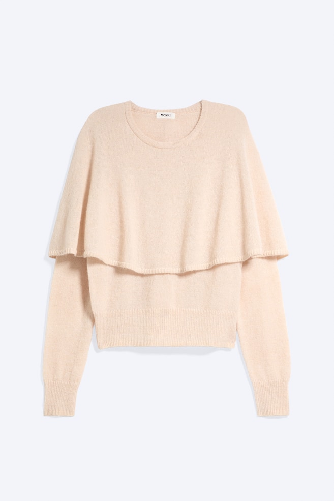 Ein hell pfirsichfarbener Strickpullover ist von vorne zu sehen, mit rundem Ausschnitt, langen Ãrmeln mit Rippbündchen und einer integrierten Cape-Ãberlage, die über die Schultern fällt. Der Pullover ist mit einem Rippbund abgeschlossen. - 1