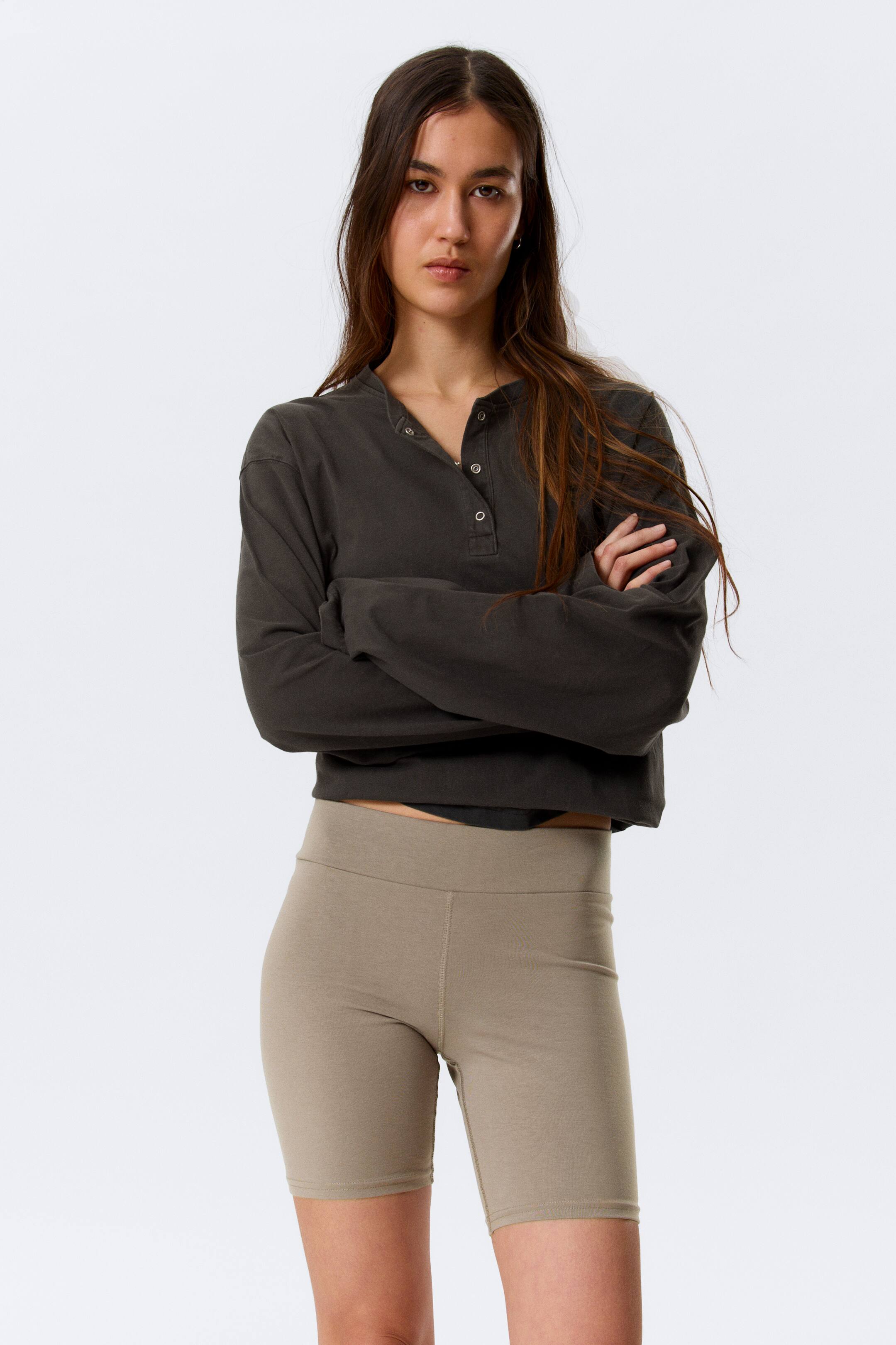 Visa större bild: Den mörkgrå långärmade henley-tröjan med kort längd har en synlig knappslå och presenteras tillsammans med ljusbruna, åtsittande cykelshorts. Shortsen har en hög midja och når till mitten av låret, med båda produkterna vända framåt.