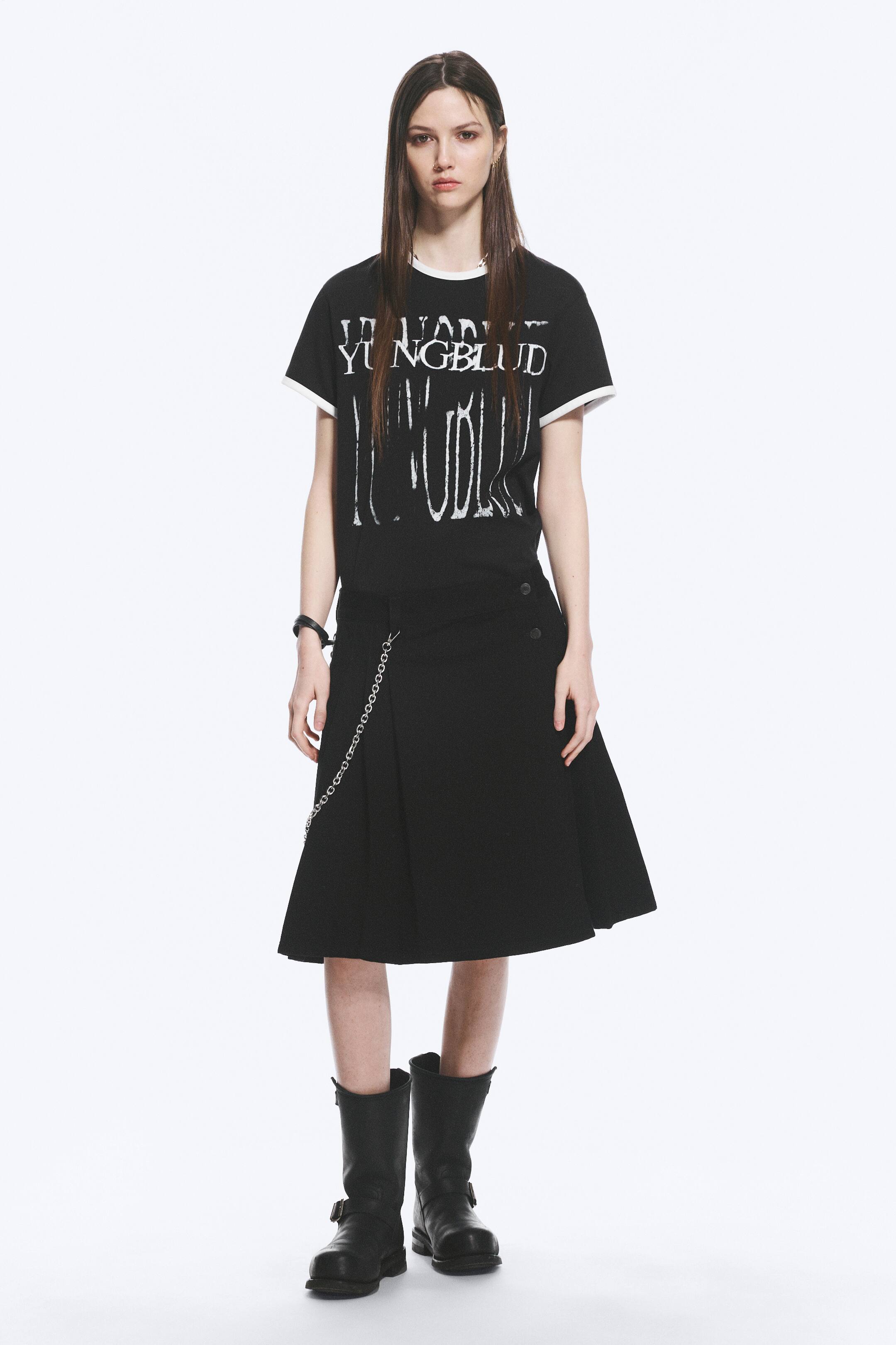 Grösseres Bild ansehen: Das Produkt ist ein schwarzes Ensemble, nach vorne gerichtet, bestehend aus einem T-Shirt mit weißem Ringerkragen und Ärmelbündchen, das abgenutzten weißen Text einschließlich "YUNGBLUD" auf der Vorderseite zeigt. Der schwarze Midirock ist mit einer Wickeloptik vorne gestaltet, gesichert durch zwei Knöpfe an der rechten Hüfte und verziert mit einer silbernen Kette, die von der linken Hüfte hängt, abgerundet durch schwarze Stiefel, die bis zur Mitte der Wade reichen und den gestylten Look vervollständigen.