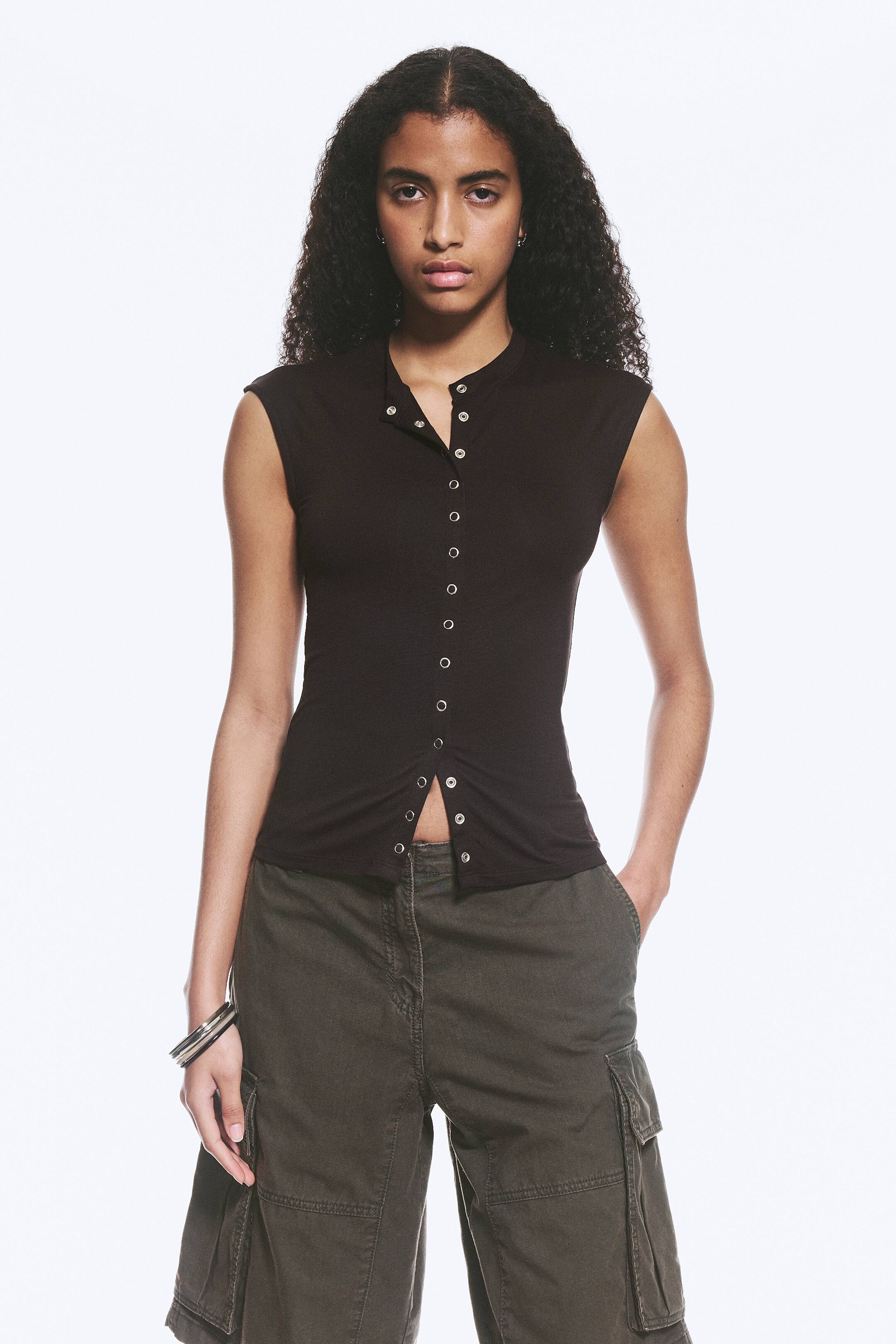 Se større billede: En mørkebrun ærmeløs top med en stand-up krave og en fuld længde stolpelukning med adskillige sølvfarvede trykknapper ned langs midten foran. Den sidder tæt til kroppen og slutter ved taljen.