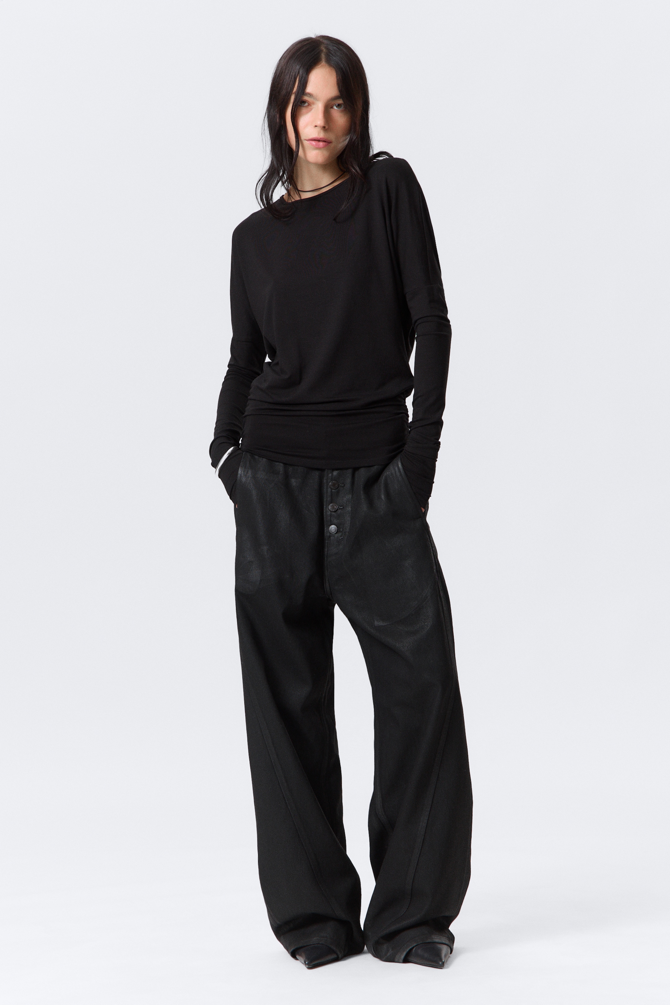 Zobacz większy obraz: Czarna bluzka z długim rękawem jest noszona, charakteryzująca się szerokim dekoltem i luźnym, drapowanym krojem, z wydłużonym dołem zebranym i warstwowym w talii. Czarnie szerokie spodnie również są noszone, z widocznym zapięciem na wiele guzików, obniżonym krokiem i teksturowanym wykończeniem, z nogawkami opadającymi na buty.