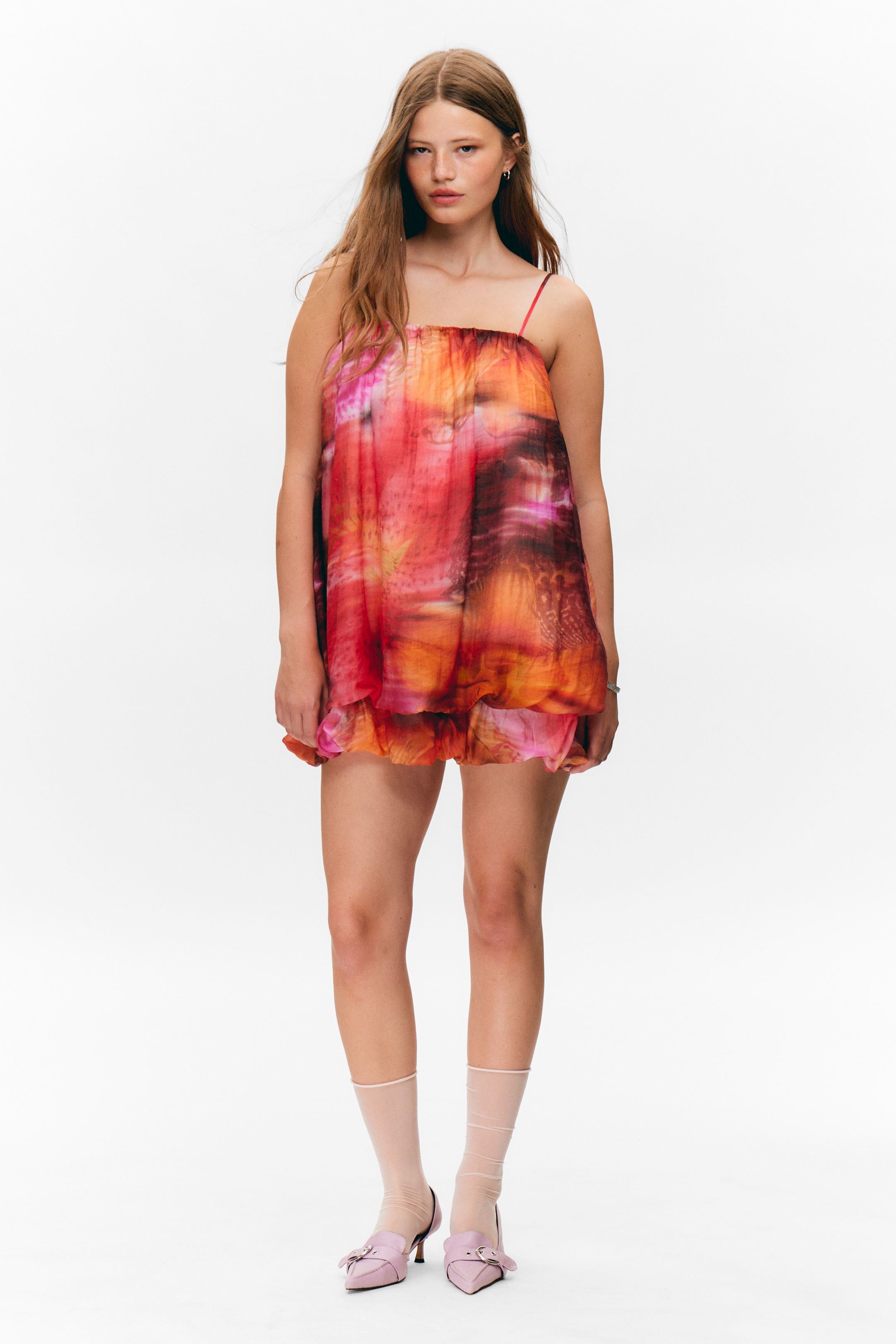 Ver imagem maior: O vestido está a ser apresentado de frente, apresentando um padrão vibrante e abstrato em tons de laranja, vermelho e rosa. É desenhado com alças finas tipo esparguete e um corpete franzido, criando uma silhueta volumosa e curta com uma bainha em camadas visível.