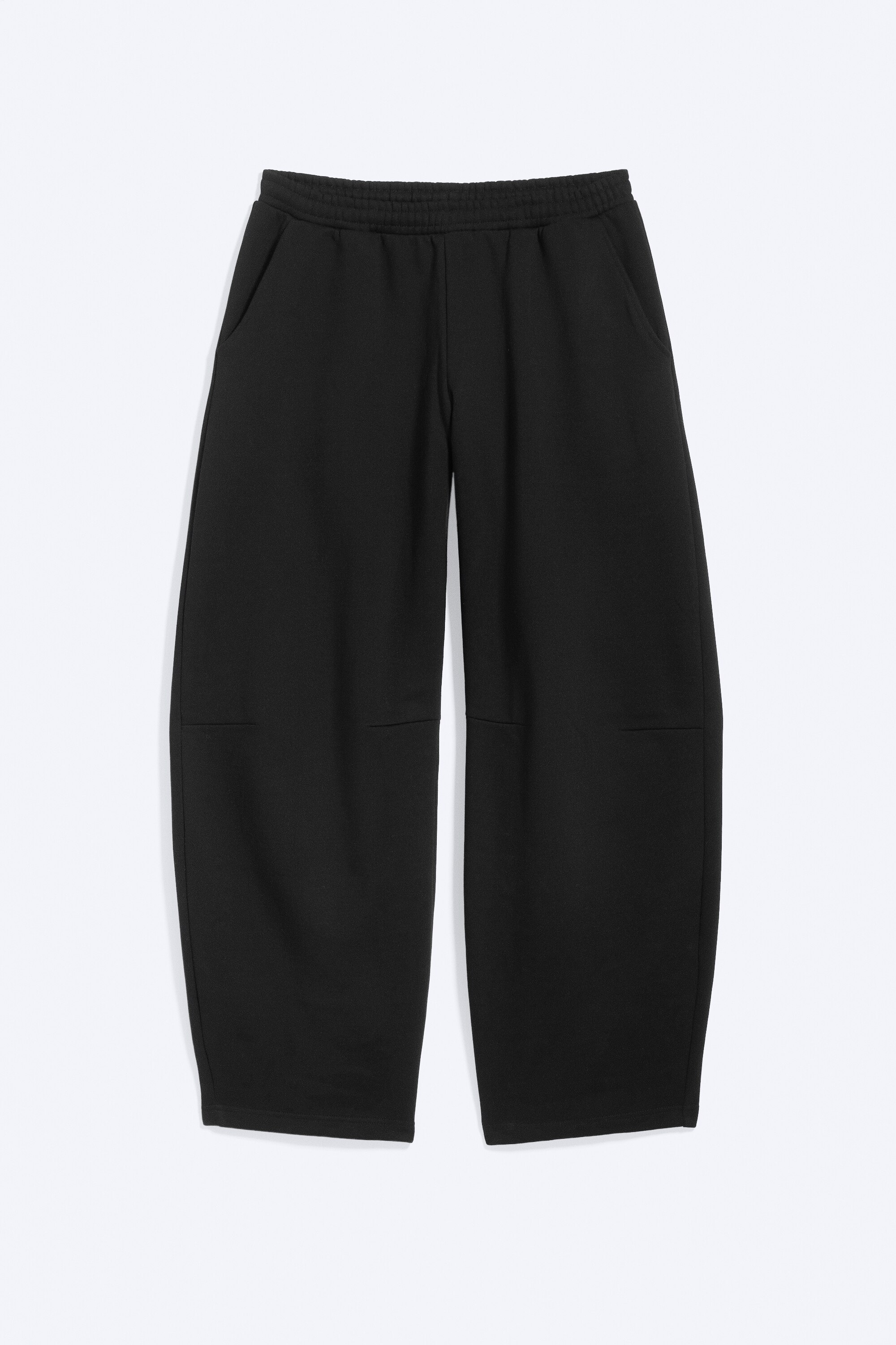 Un pantalon de jogging noir Ã  coupe ample, dotÃ© dâune taille Ã©lastiquÃ©e et dâune jambe arrondie de type barrel, est portÃ© par le modÃ¨le. Le pantalon est associÃ© Ã  un hoodie court noir zippÃ© assorti, Ã  un t-shirt blanc et Ã  des sneakers noires.