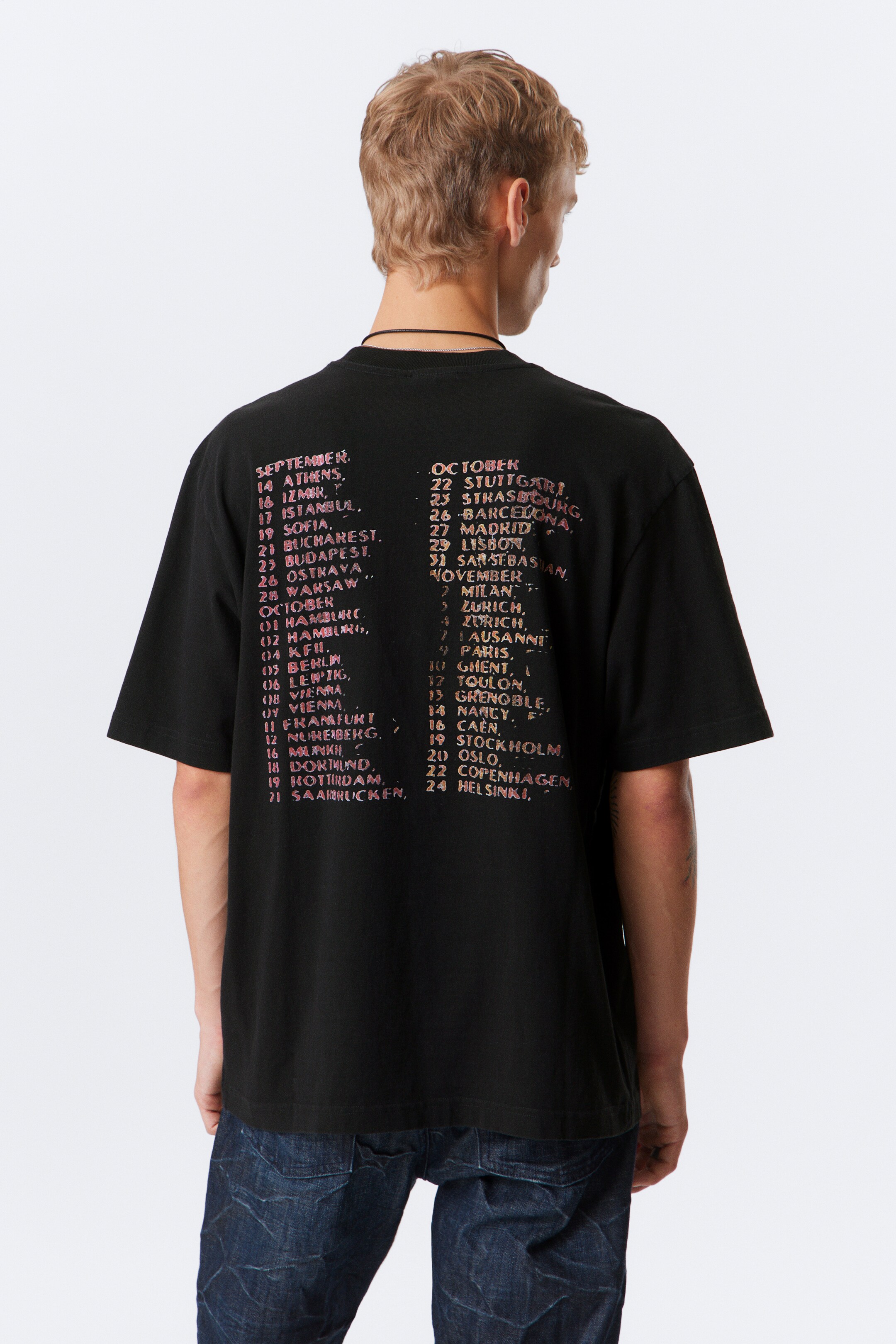 Vezi imaginea mai mare: Un tricou negru este prezentat din spate, având un imprimeu grafic deteriorat pe partea superioară a spatelui care listează orașe și date într-o culoare roz și portocaliu șters. Este proiectat cu un guler rotund și mâneci scurte, prezentând o croială lejeră.