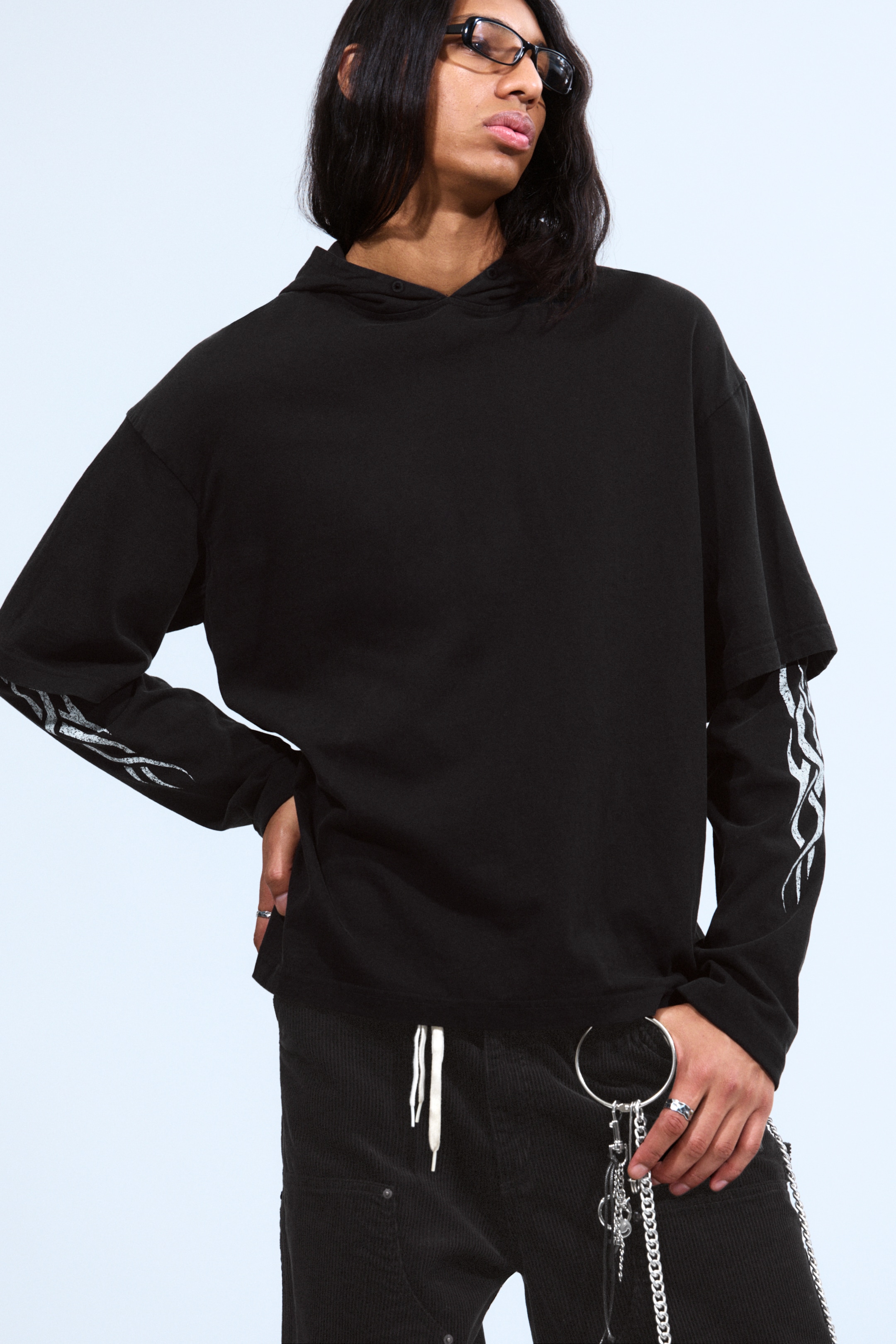 View larger image: A black oversized hoodie is being presented, with its short sleeves revealing a black long-sleeve tee underneath. The long-sleeve tee is featuring stylized silver flame graphics on both arms.