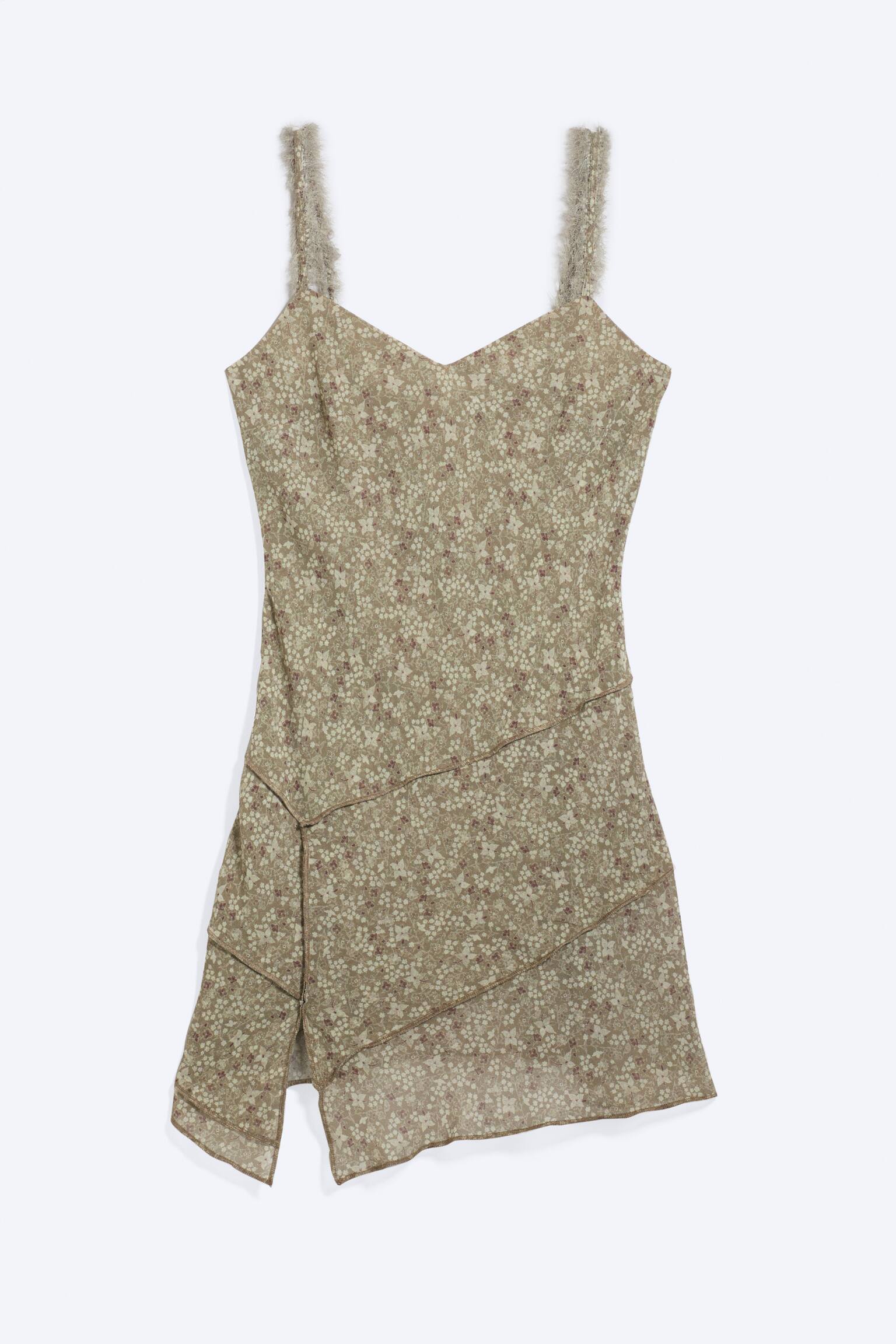 O rochie tip slip cu imprimeu floral este prezentată din față, având un fundal verde pal cu un model dens de flori mici crem și maro-roșcat. Este proiectată cu un decolteu în V, bretele subțiri cu franjuri de culoare deschisă și un tiv asimetric creat prin cusături diagonale care formează panouri stratificate, semi-transparente.