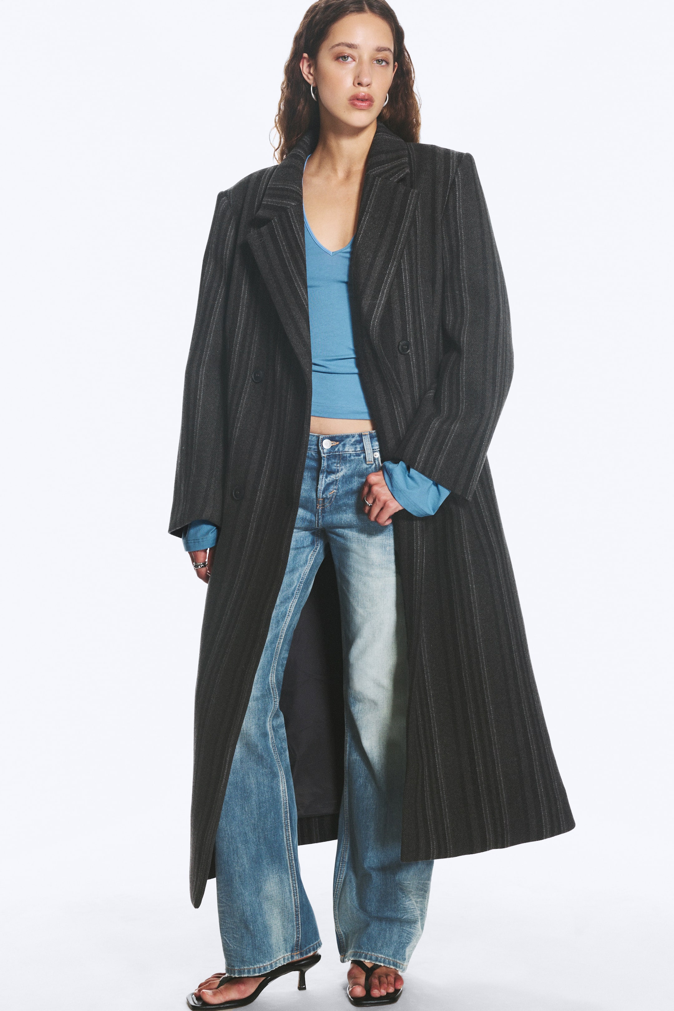 Agrandir l'image: Le manteau long à fines rayures gris foncé présente des revers crantés et une fermeture croisée à double boutonnage avec deux boutons visibles. Il est porté ouvert, mettant en valeur sa silhouette longue et fluide ainsi qu’un haut bleu à manches longues et un jean en dessous.