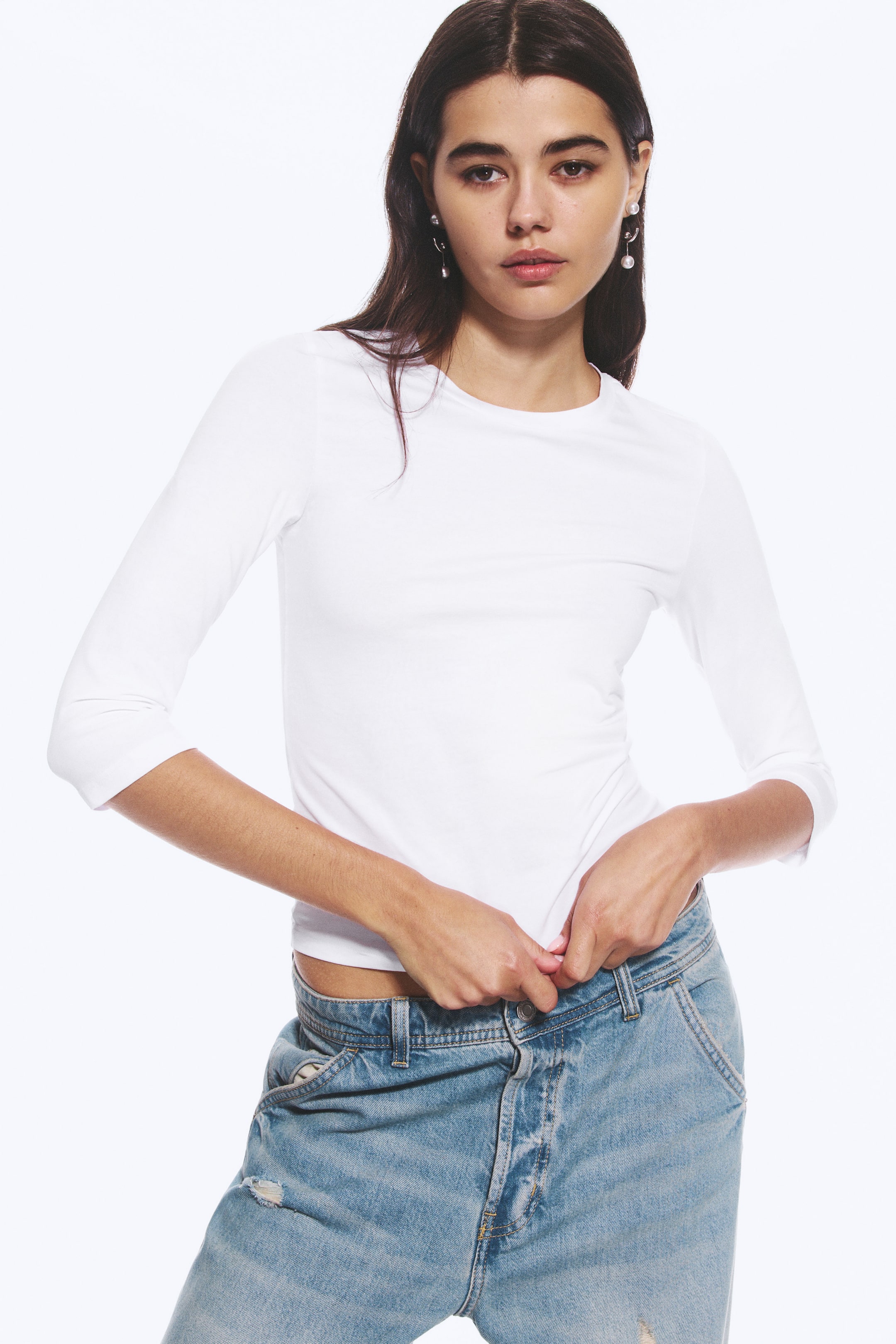 Grösseres Bild ansehen: Das Model trägt ein weisses Oberteil mit Rundhalsausschnitt und Dreiviertelärmeln und hält den Saum am Bund einer hellblauen Jeans.