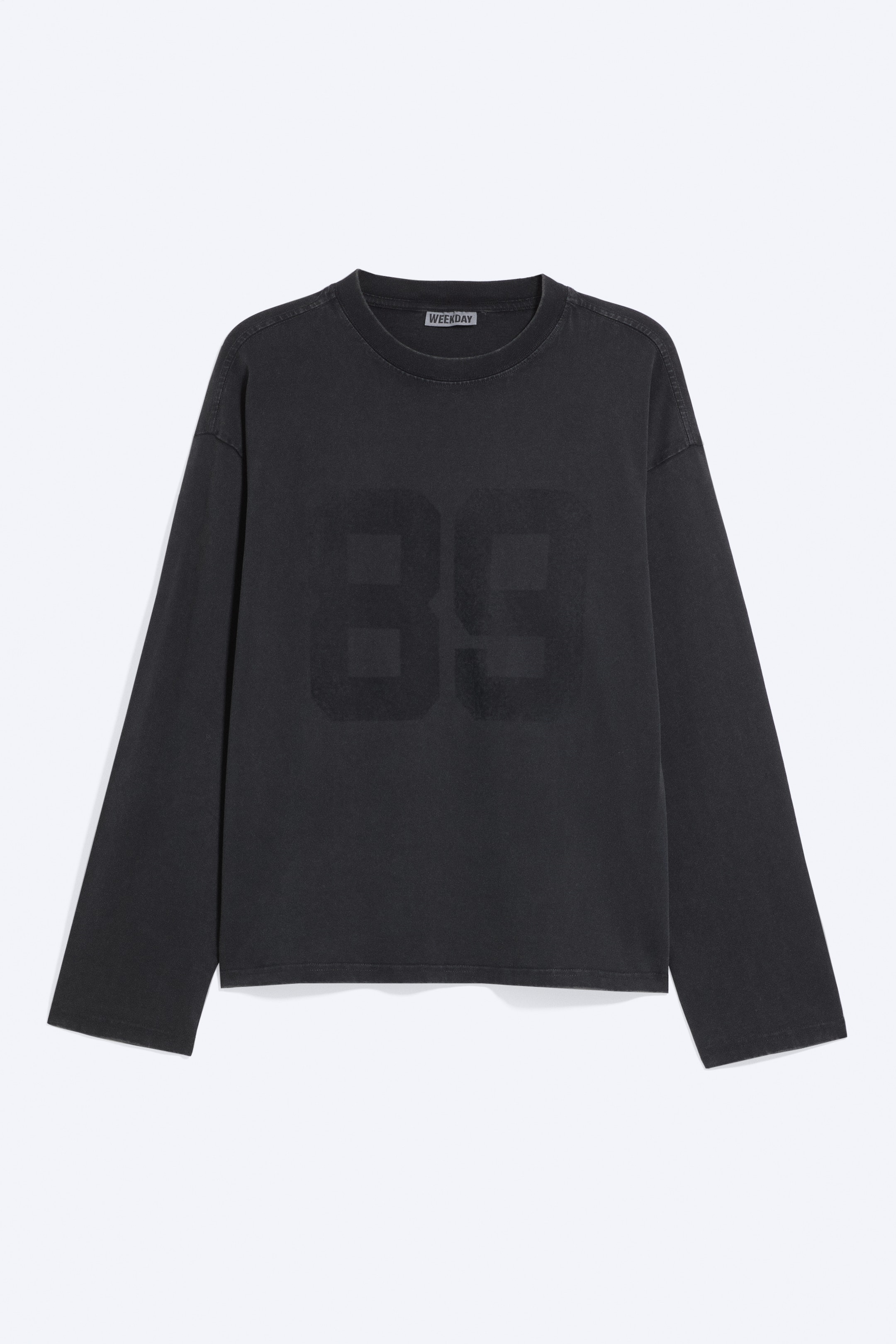 Se større billede: En boxy T-shirt med lange ærmer i falmet sort vises forfra og har rund hals i ribstrik, sænkede skuldre og et stort, tonalt "89"-motiv trykt på brystet.