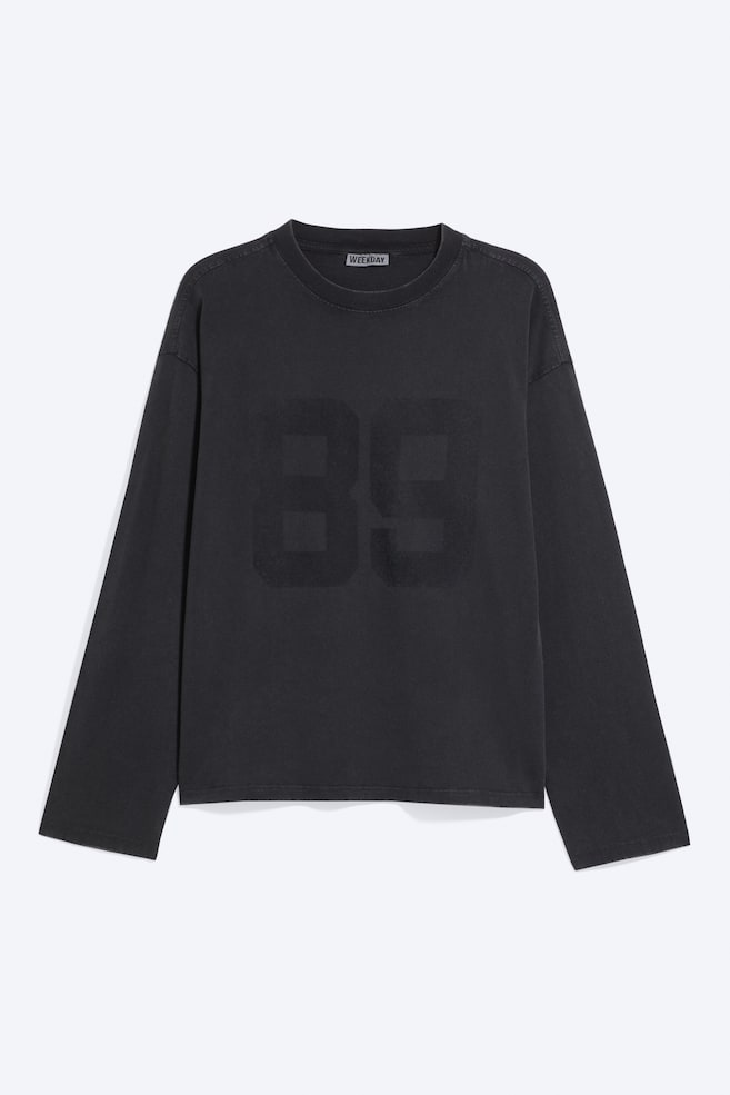 En boxy T-shirt med lange ærmer i falmet sort vises forfra og har rund hals i ribstrik, sænkede skuldre og et stort, tonalt "89"-motiv trykt på brystet. - 1
