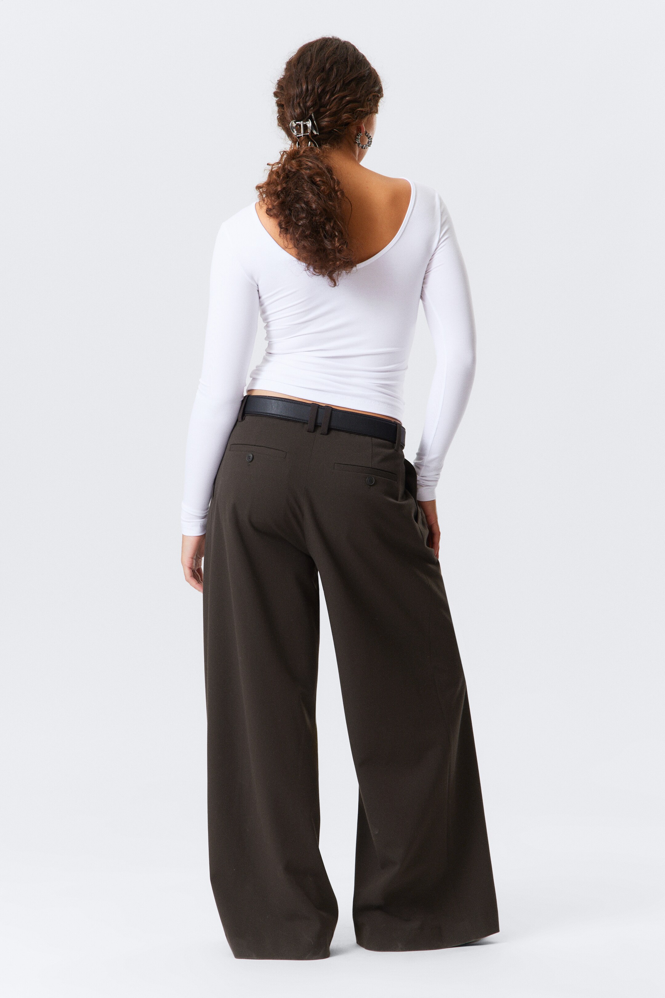 Visualizza immagine più grande: Un top bianco aderente a maniche lunghe con un ampio scollo posteriore è abbinato a pantaloni marrone scuro a gamba larga, visto da dietro. I pantaloni presentano una cintura nera infilata nei passanti e due tasche posteriori con bottoni visibili.