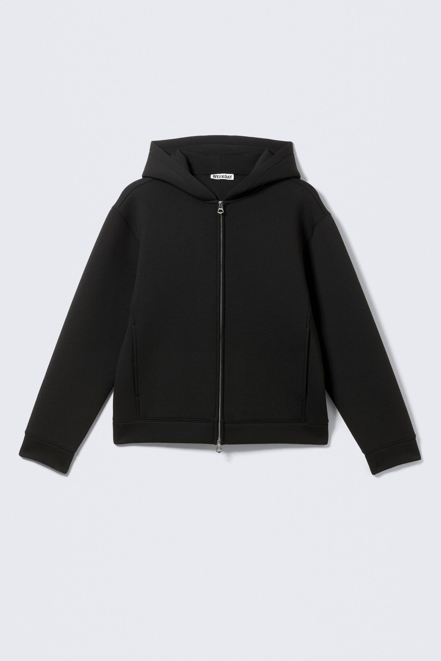Un sweat à capuche noir à fermeture éclair est présenté de face, avec une fermeture éclair argentée qui descend au centre et une capuche intégrée. Le sweat présente des manches longues avec des poignets simples, un ourlet droit et deux poches fendues verticales à l'avant. Une étiquette "WEEKDAY" blanche est visible à l'intérieur de l'encolure arrière.
