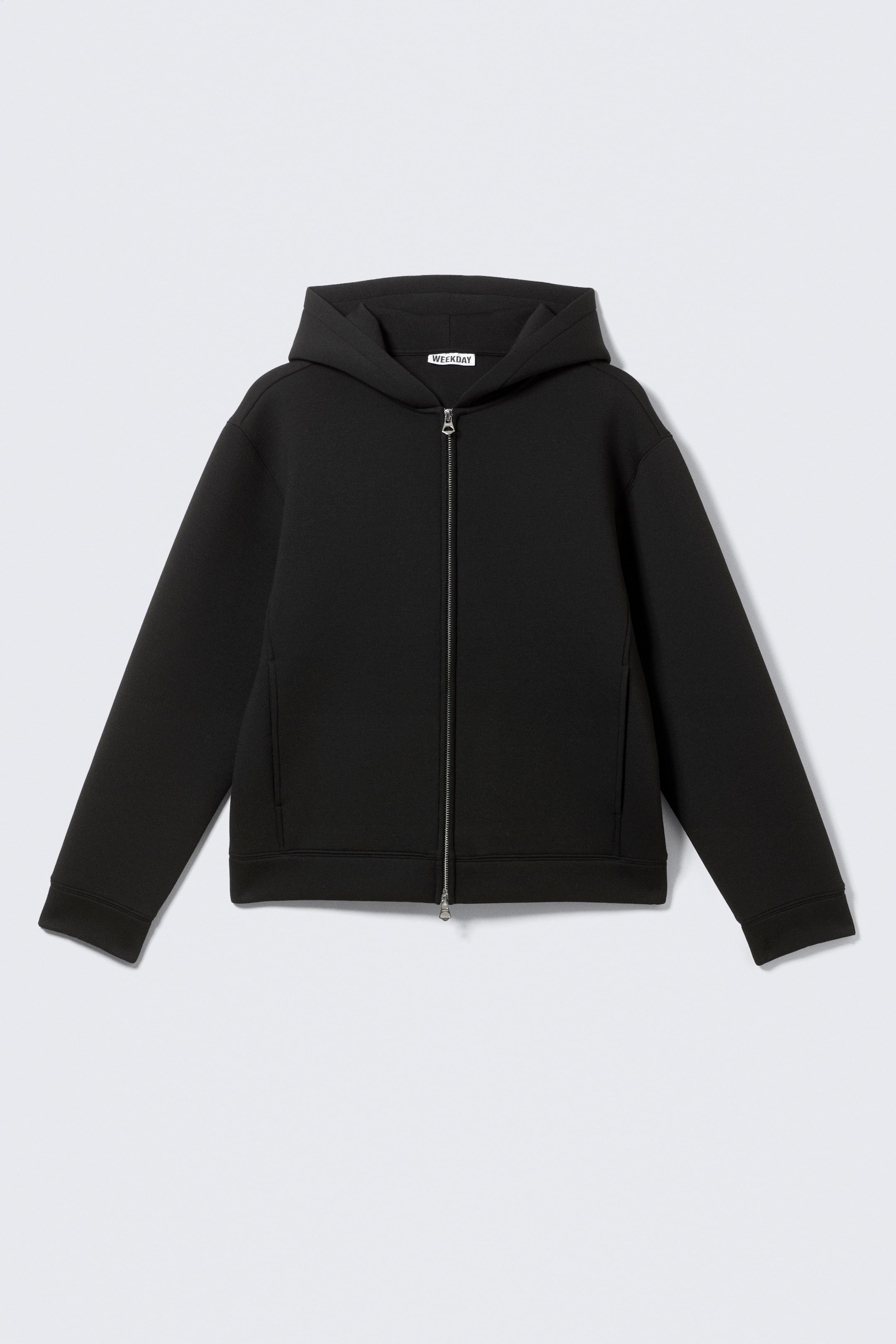 Agrandir l'image: Un sweat à capuche noir à fermeture éclair complète présente sa vue de face, avec une fermeture éclair argentée courant le long du centre et une capuche intégrée. Le sweat à capuche montre des manches longues avec des poignets simples, un ourlet droit, et deux poches fendues verticales à l'avant. Une étiquette "WEEKDAY" blanche est visible à l'intérieur de l'encolure arrière.