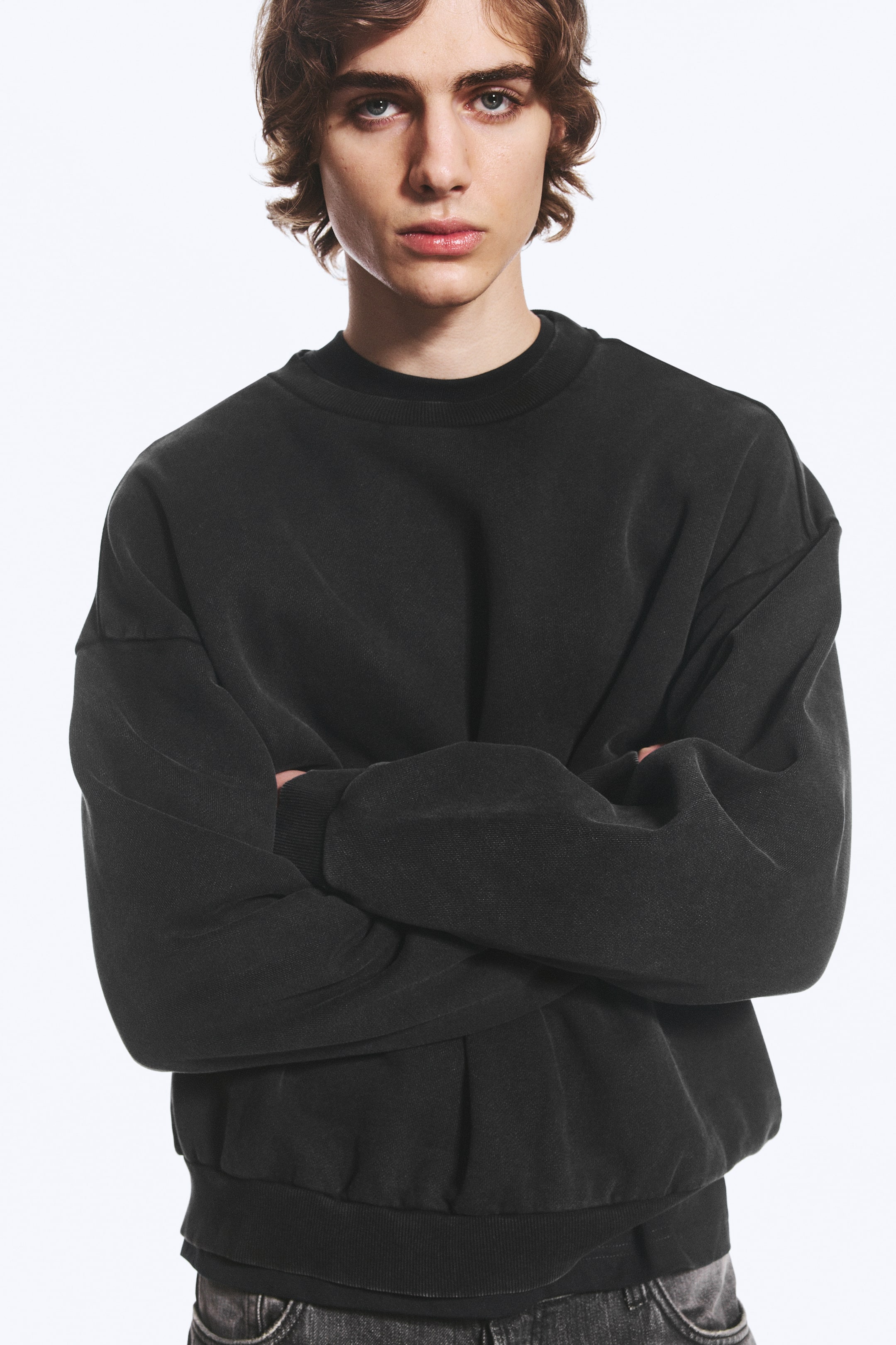Größeres Bild ansehen: Das Model trägt einen locker geschnittenen, verwaschenen schwarzen Sweatshirt und posiert mit verschränkten Armen. Das Sweatshirt verfügt über einen gerippten Rundhalsausschnitt, überschnittene Schultern und einen gerafften, gerippten Saum.