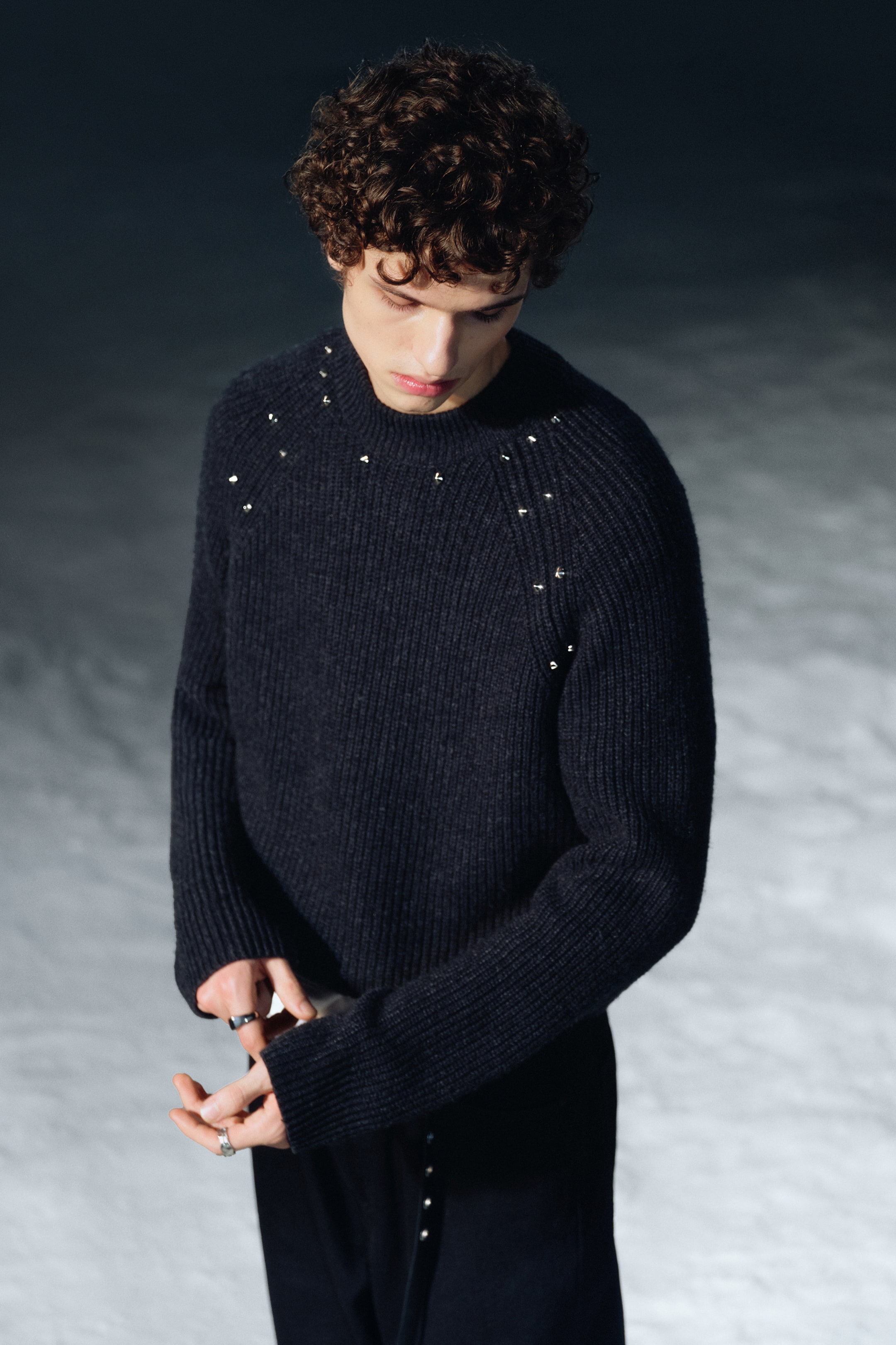 Visualizza immagine più grande: Un maglione girocollo a maglia scura e a coste viene visto da un'angolazione leggermente elevata e frontale. Presenta piccoli borchie metalliche lungo le cuciture raglan e lo scollo, e mostra maniche lunghe con polsini a coste.