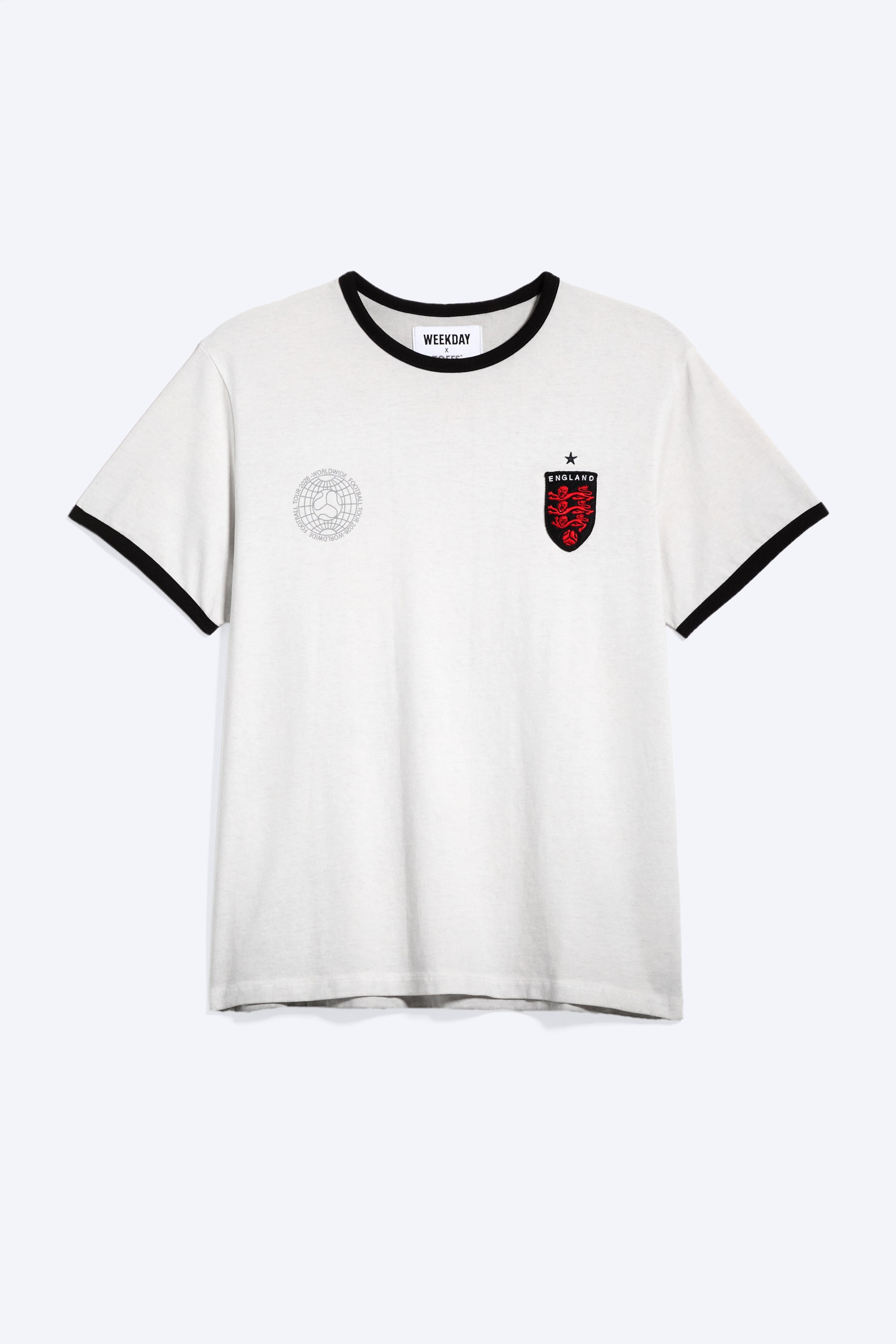 Ver imagen más grande: Una camiseta blanca roto se presenta en vista frontal, con un cuello redondo acanalado en negro y un ribete acanalado negro en los puños de sus mangas cortas. Un parche bordado en forma de escudo con tres leones rojos, el texto 'ENGLAND' y una estrella aparece en el lado izquierdo del pecho, mientras que un gráfico circular 'WORLDWIDE FOOTBALL TOUR' con un globo terráqueo estilizado es visible en el área del pecho derecho.