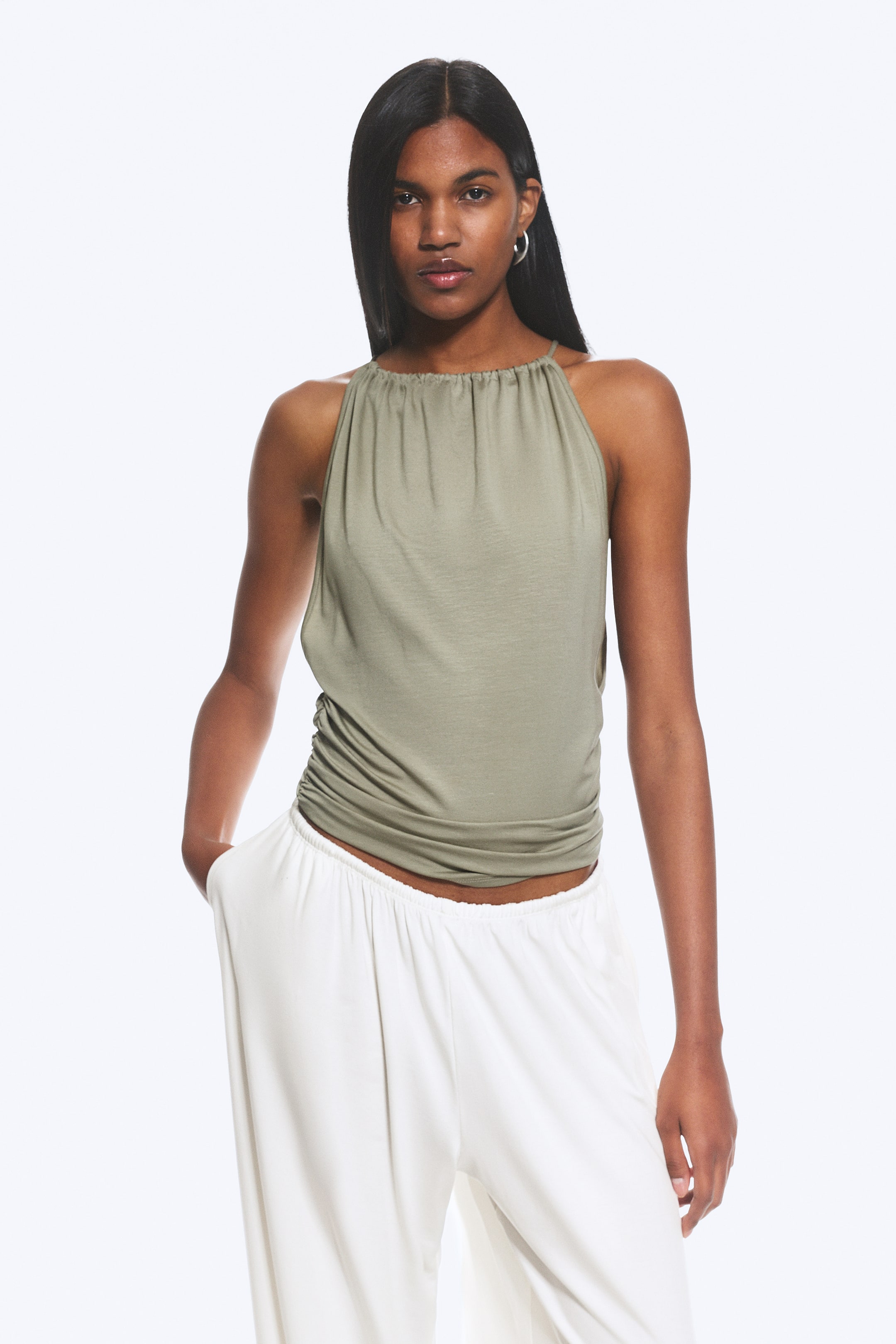 Ver imagem maior: Um top verde-azeitona com decote halter apresenta uma gola franzida com alças finas. Cai de forma solta, com franzidos visíveis ao longo do seu lado esquerdo.