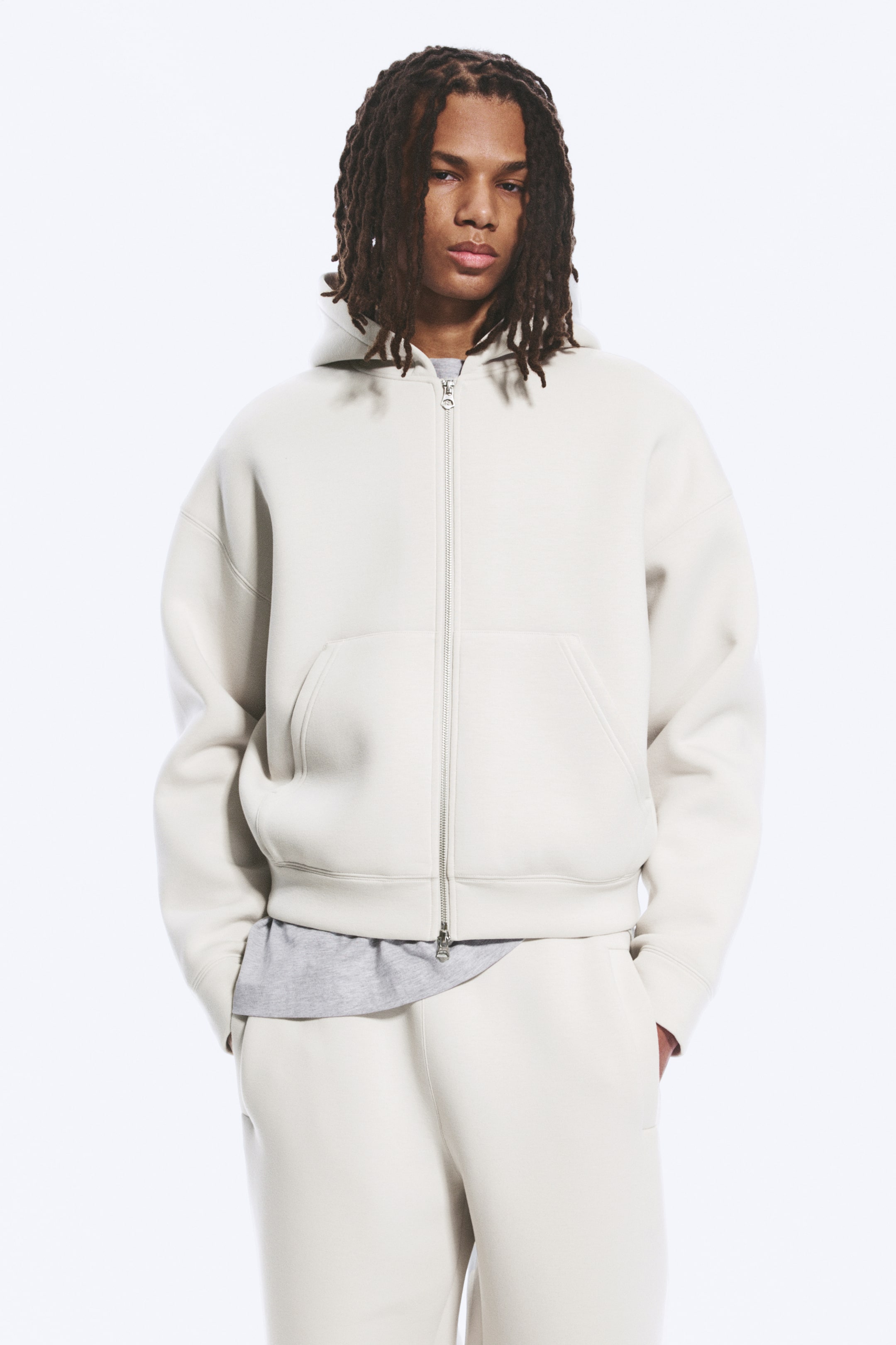 Näytä suurempi kuva: Vaaleanbeige vetoketjullinen huppari on kuvattu edestäpäin, esittäen rennon istuvuuden ja hopeanvärisen vetoketjun edessä, hupun sekä jaetun kengurutaskun. Siinä on ribatut hihansuut ja ribattu helma, ja se on puettu harmaan t-paidan päälle.