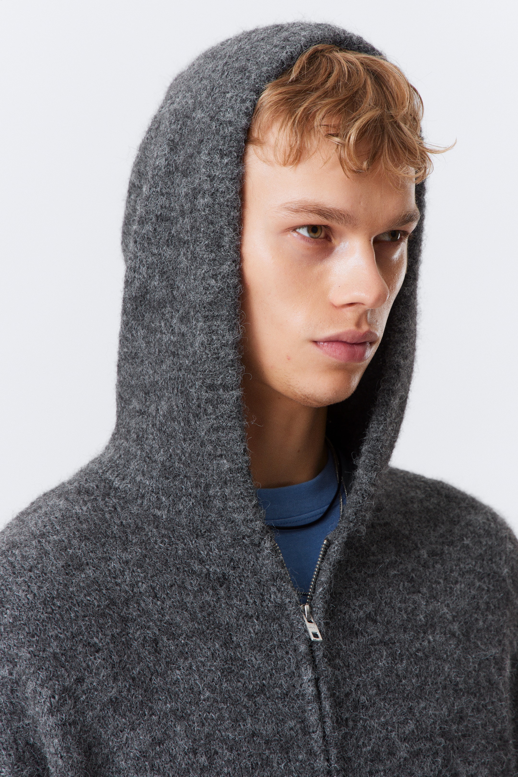 View larger image: The upper part of a dark gray fuzzy knit zip-up hoodie is being shown, with its hood up. A silver-toned zipper is visible down the front, partially unzipped over a blue crew-neck top.