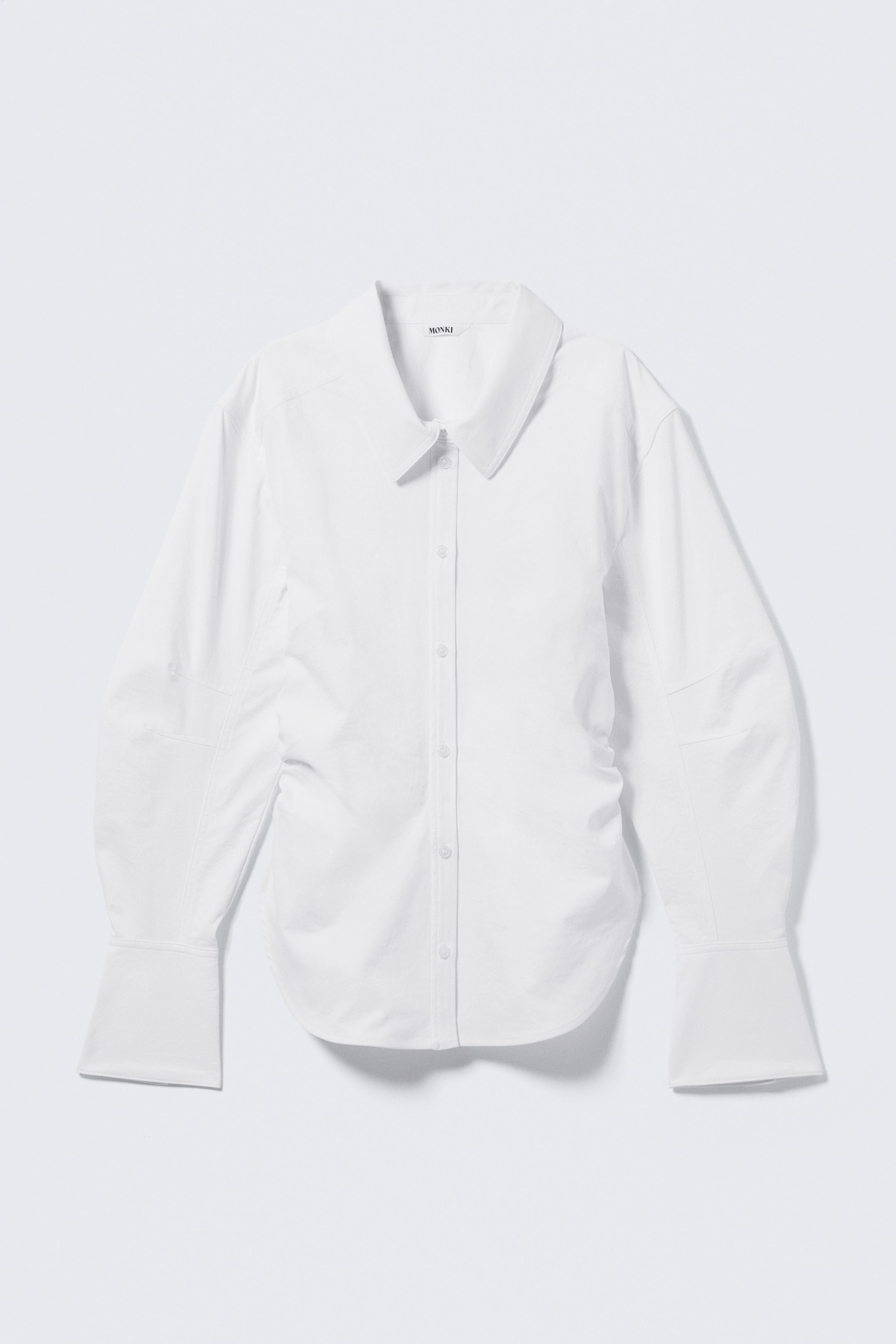 Visualizza l'immagine ingrandita: Una camicia bianca con bottoni è presentata in vista frontale, caratterizzata da un colletto a punta e un'abbottonatura completa con bottoni bianchi. Presenta maniche lunghe con polsini ampi ripiegati, dettagli di arricciatura lungo le cuciture laterali e le maniche interne, e pannelli strutturati sulla parte superiore delle maniche esterne.